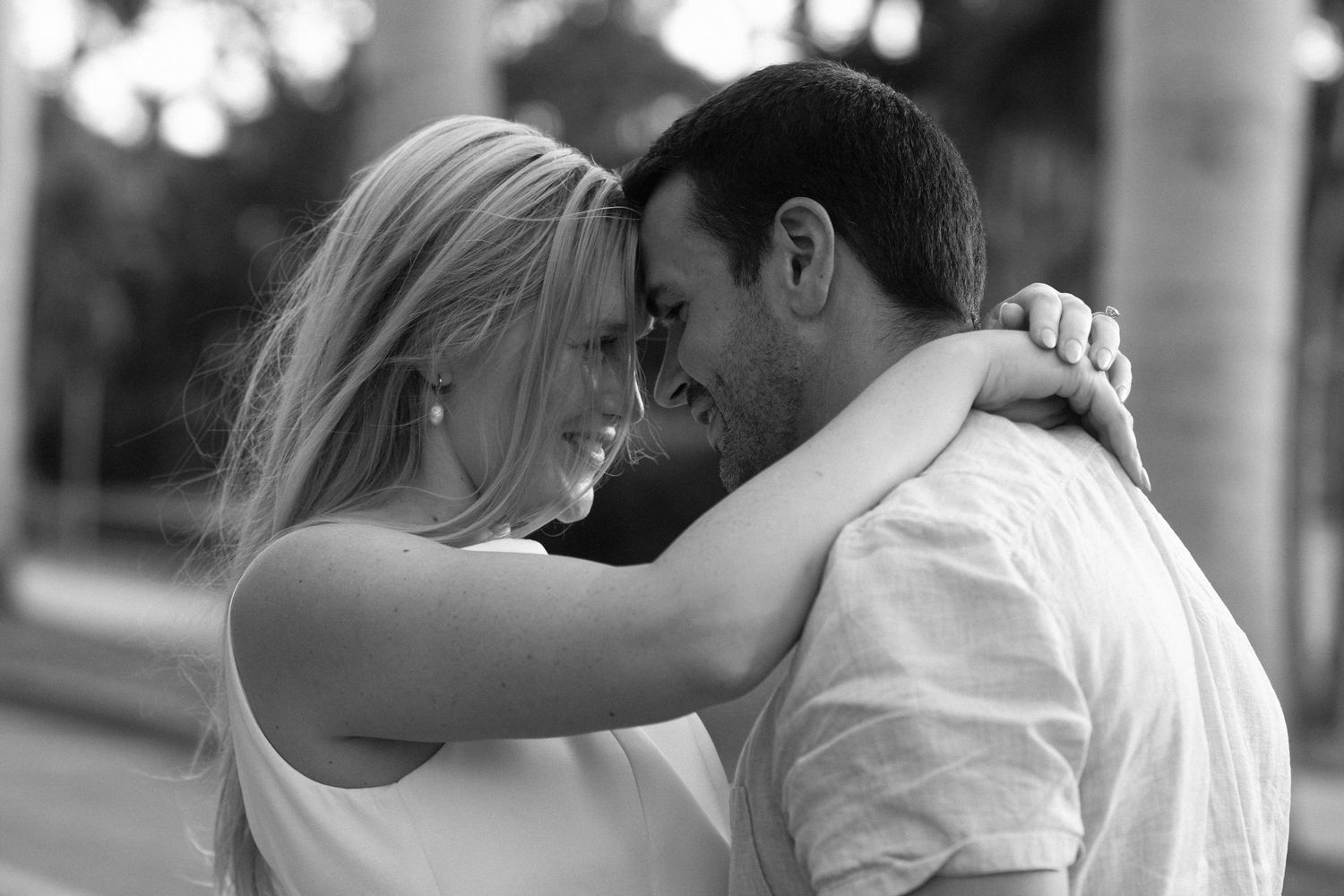 Black and white photo of couple embracing between white columns with soft romantic lighting.