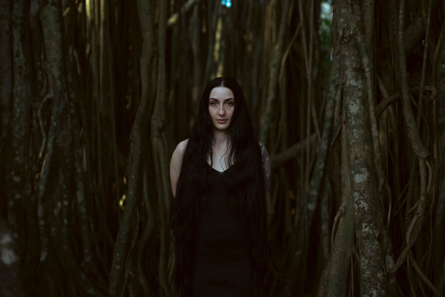 A figure in dark gothic attire stands among shadowy trees in a moody forest setting.