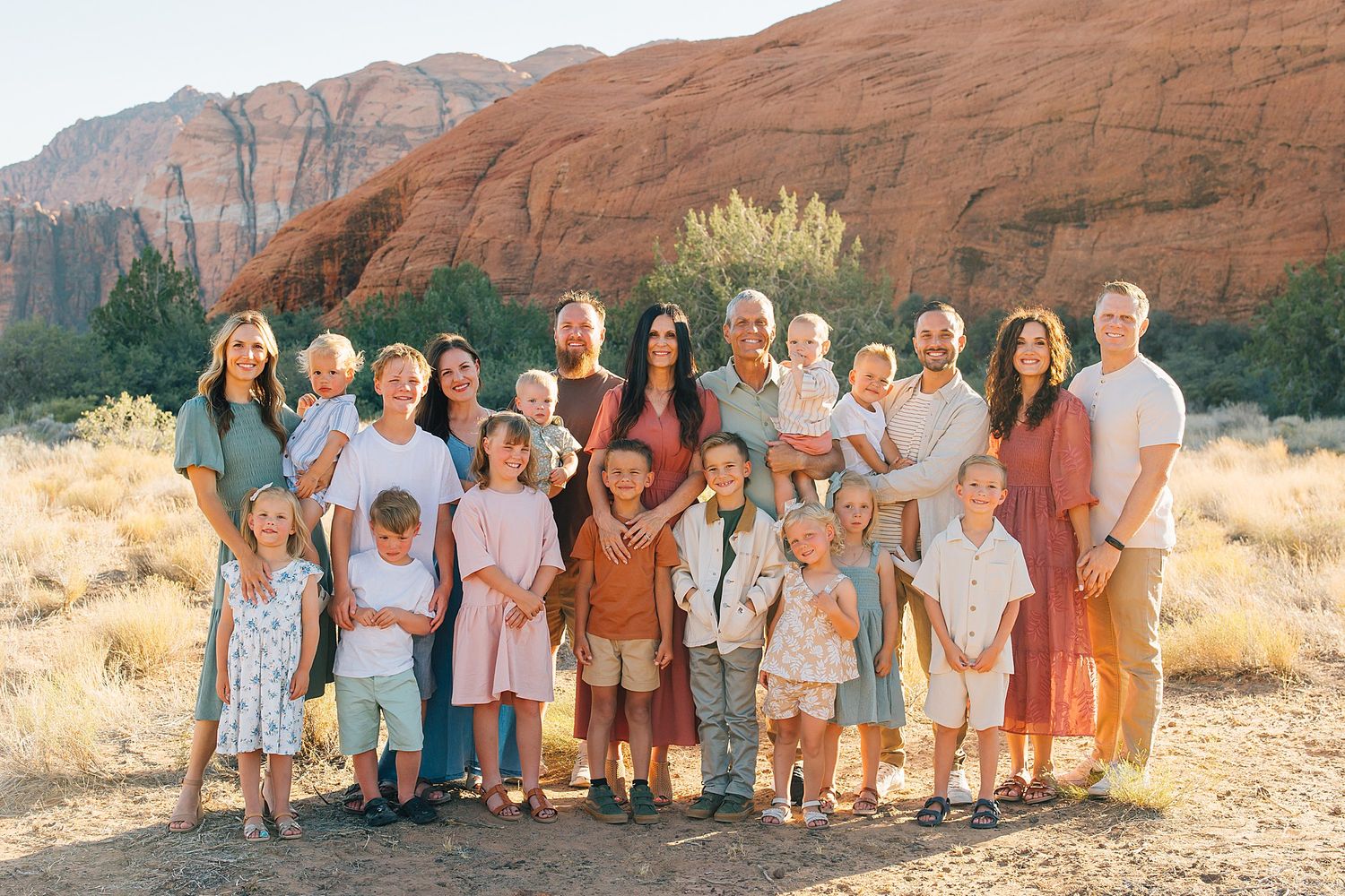Large group in coordinated earth-tone outfits pose together against stunning red rock formations in desert landscape.