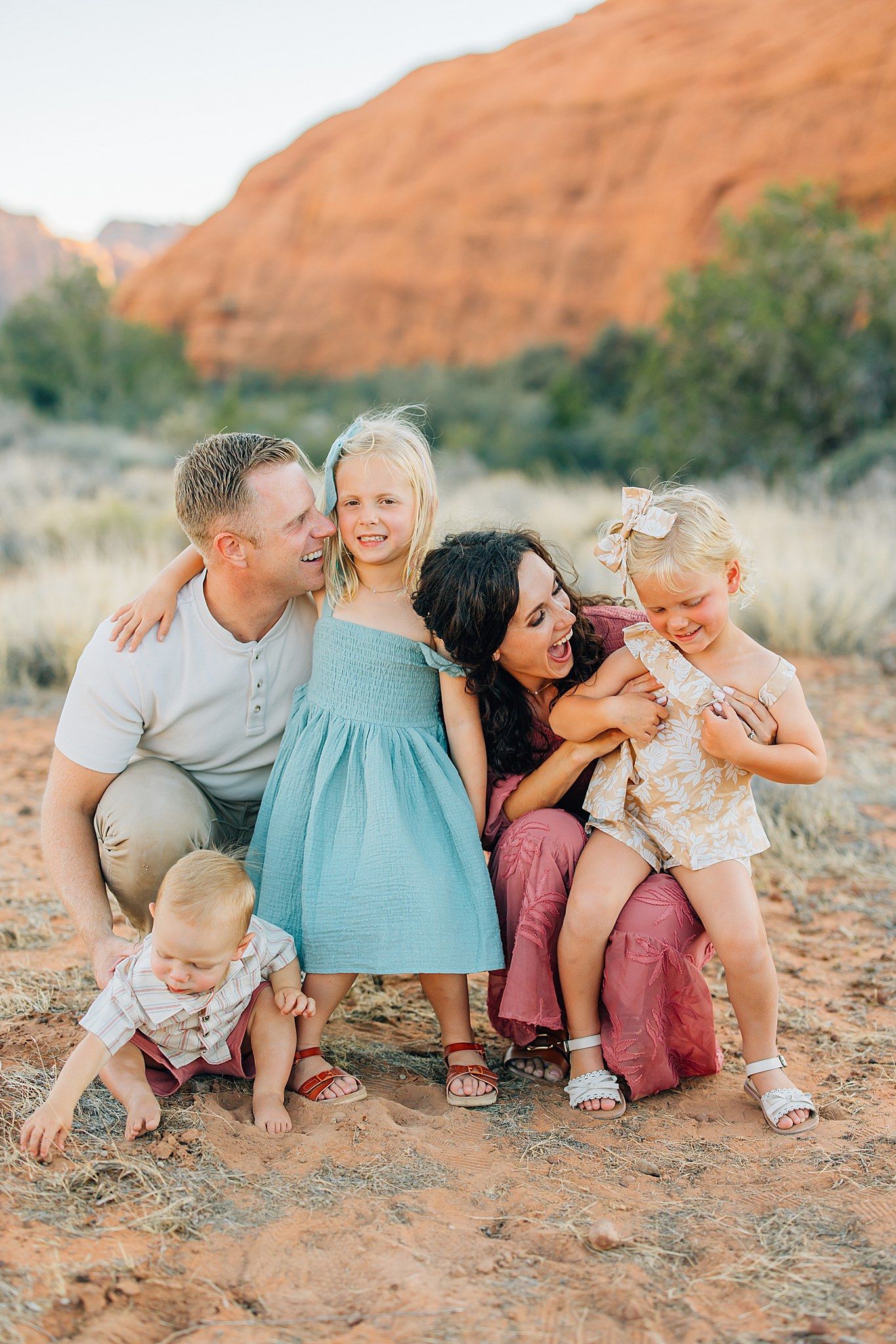 A family shares joyful moments together while sitting on desert ground with red cliffs in background.