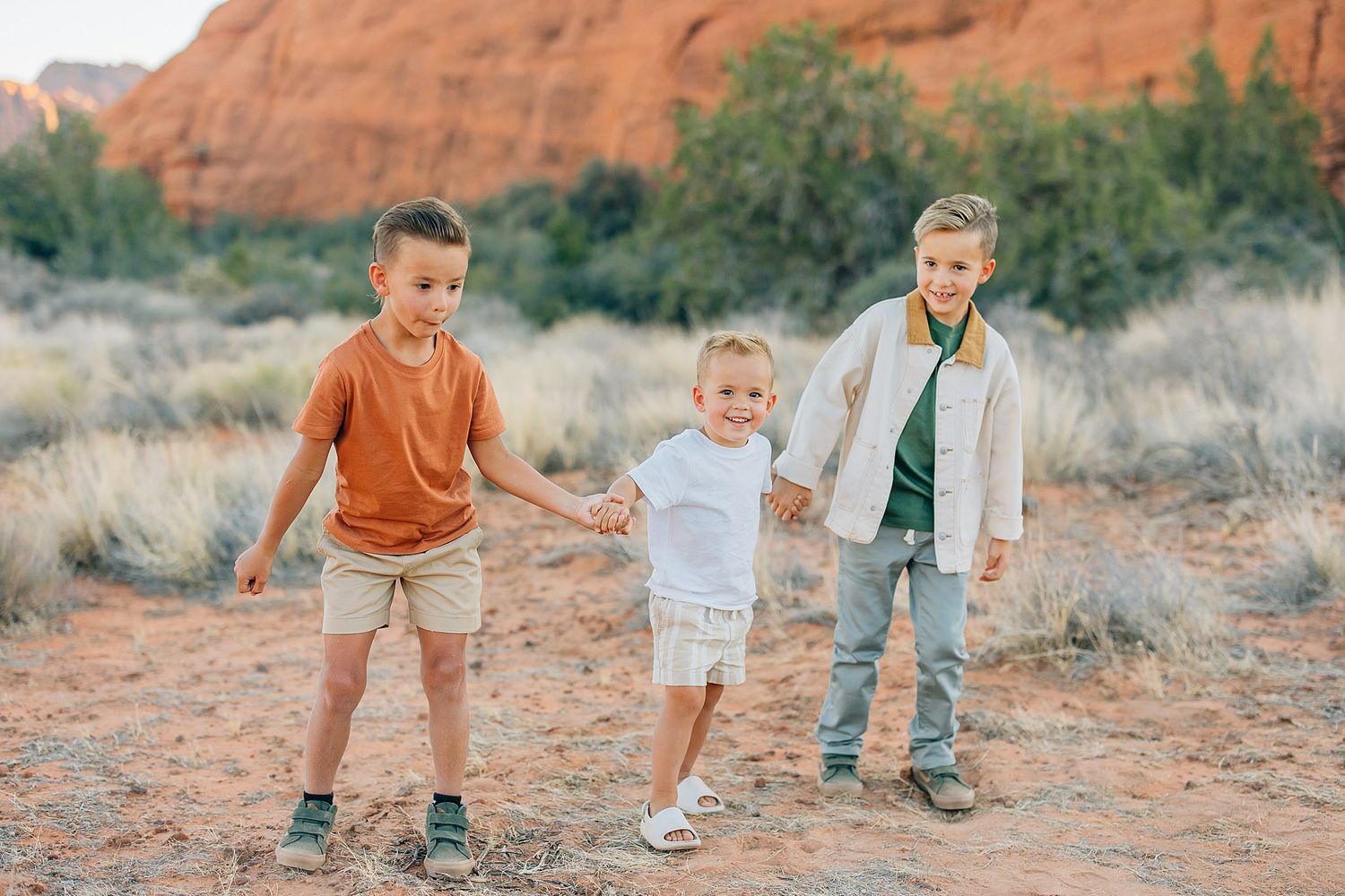 Three children playing and holding hands against backdrop of red desert cliffs and brush.