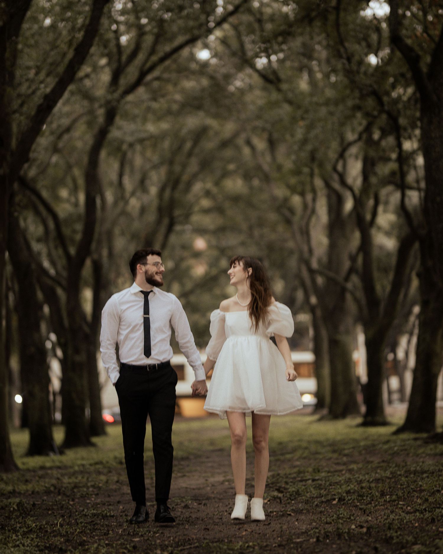 Playful couple walks through tree-lined path in formal attire.
