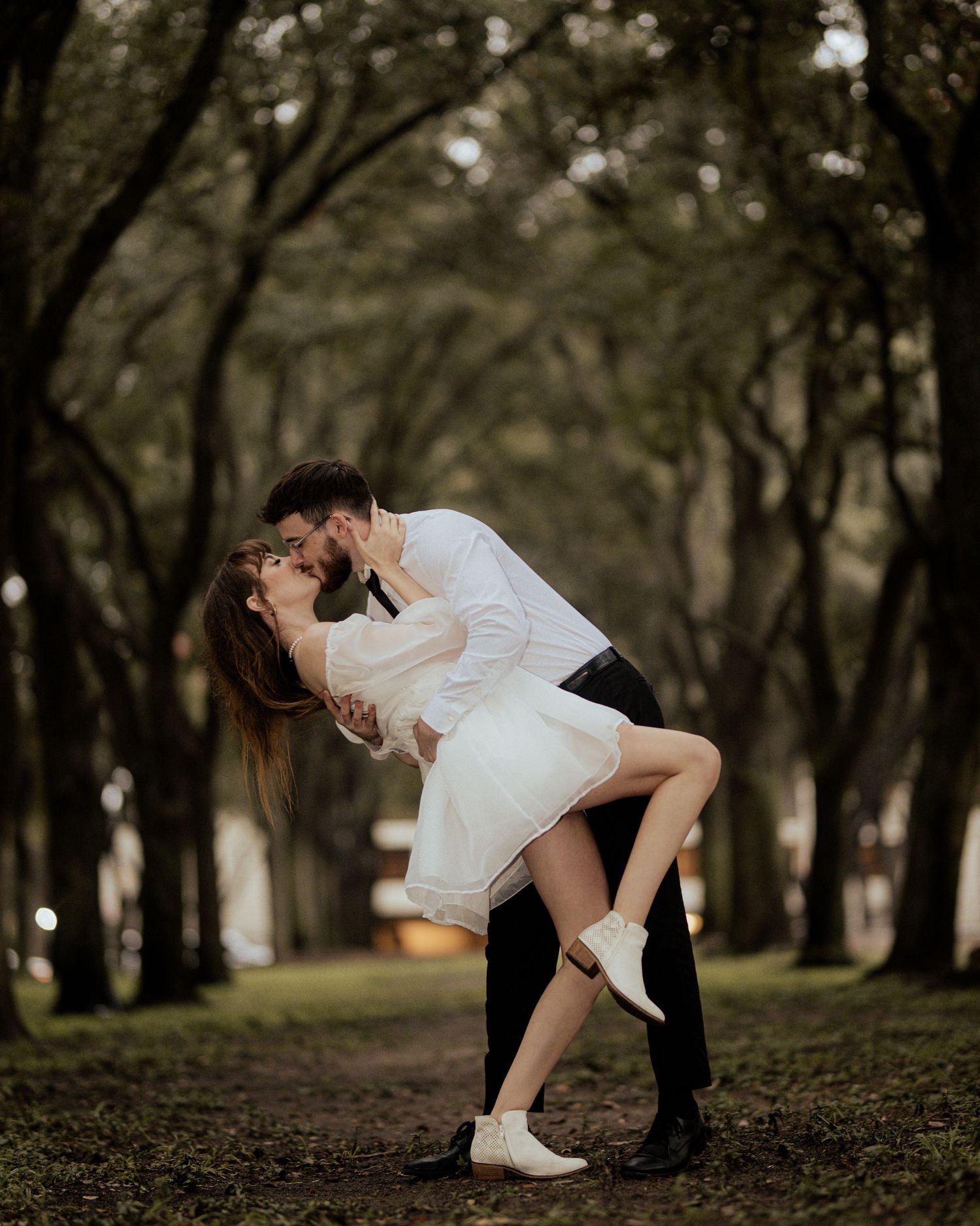 Romantic couple shares intimate moments in a park setting with tree-lined backdrop.