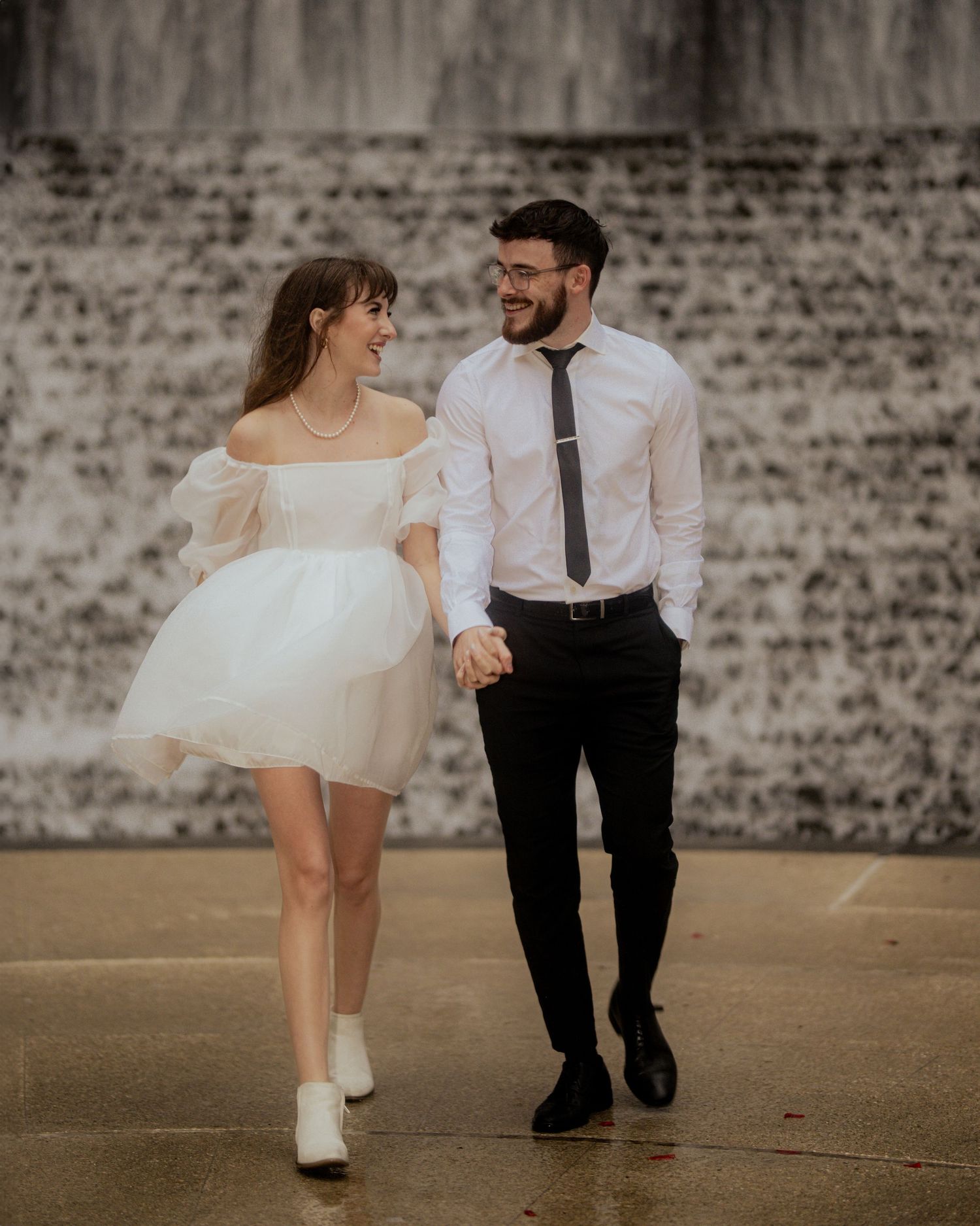Couple in formal wear dances playfully against textured brick wall background.