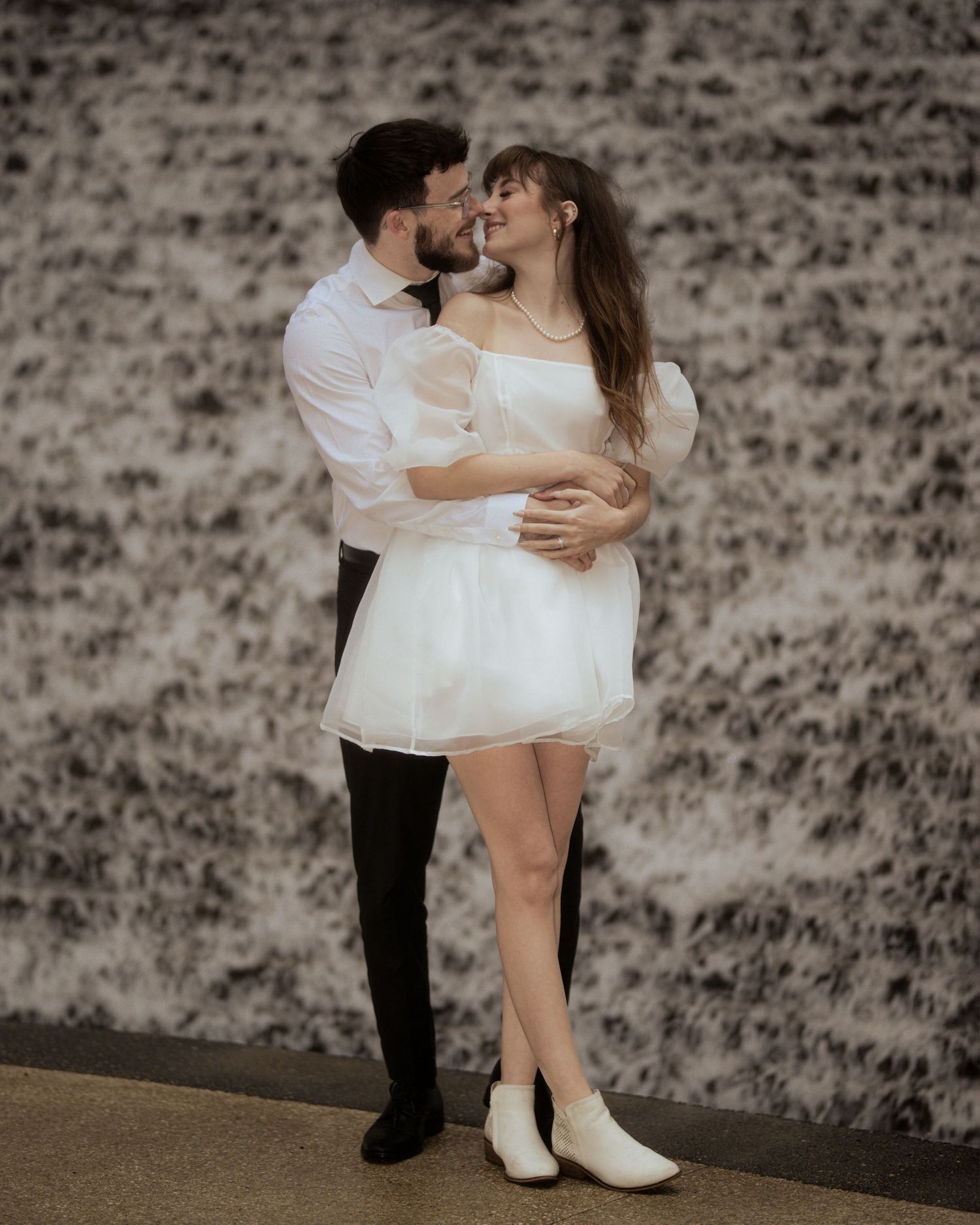 A couple in white and black formal wear embrace while dancing together against a stone wall backdrop.