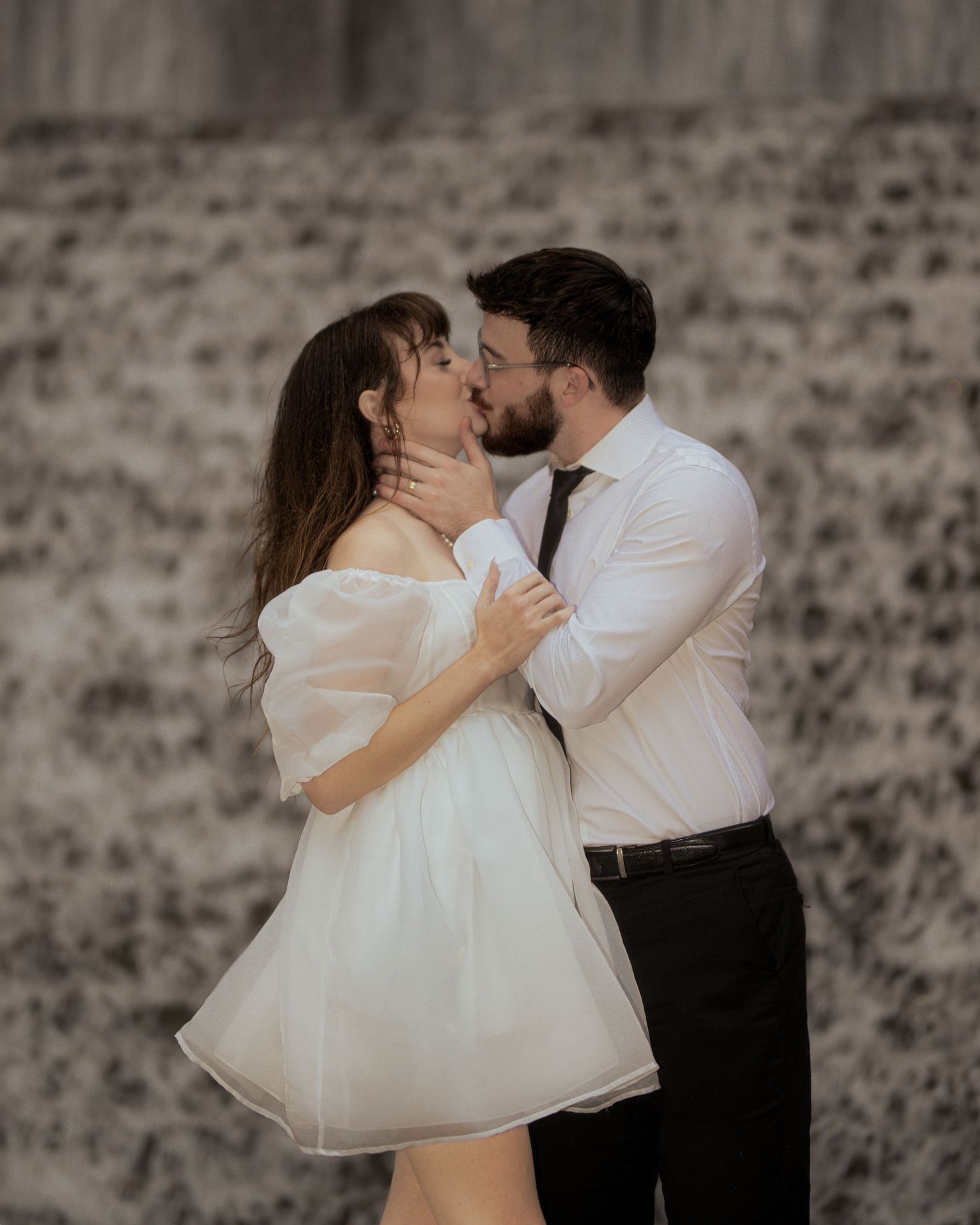 A couple shares romantic dance poses in formal attire against a rustic stone wall background.