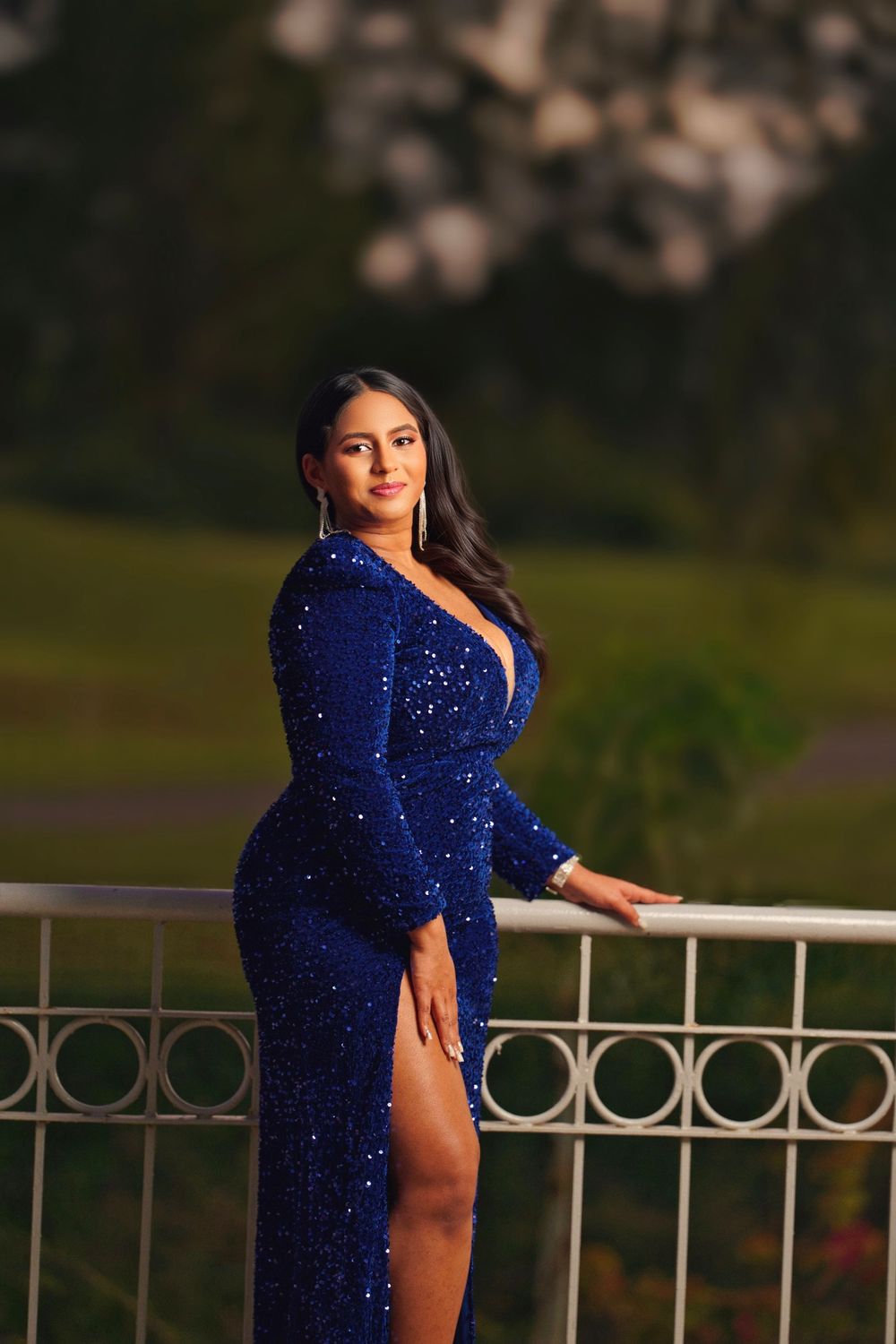A woman poses on a balcony at sunset wearing a sparkling royal blue evening gown with a thigh-high slit.