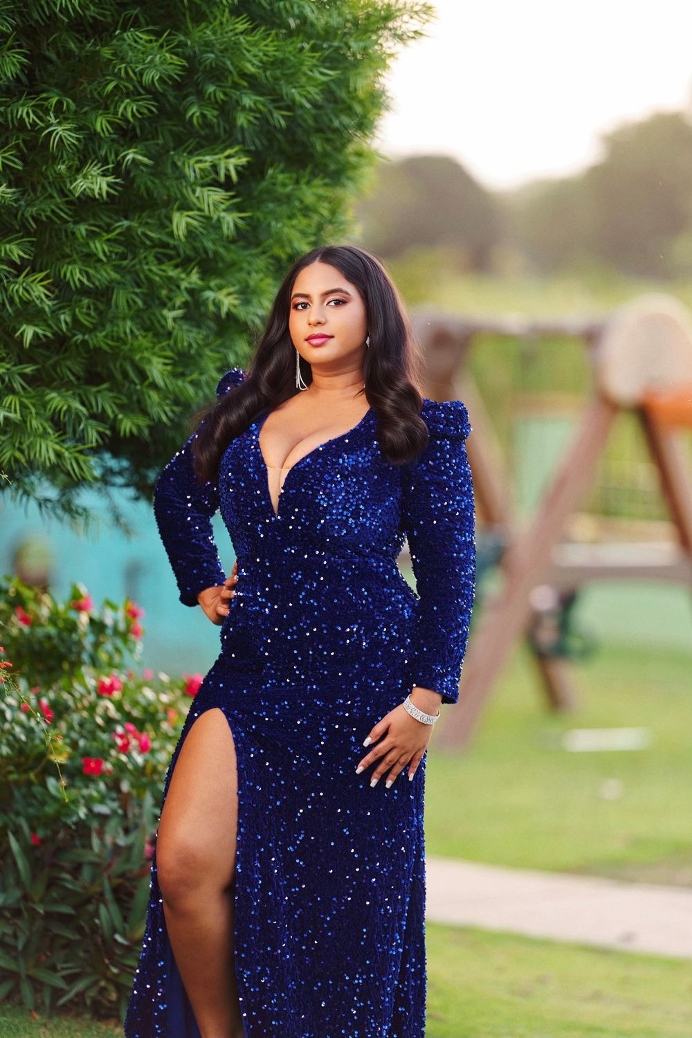 A woman poses outdoors in a sparkling royal blue velvet dress with long sleeves against a green garden background.