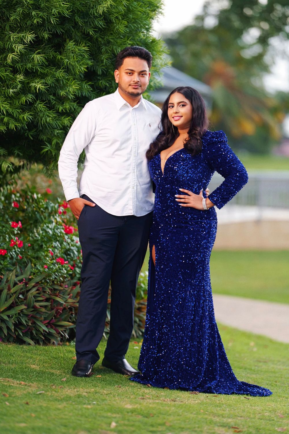 A couple poses outdoors in formal attire with the person in a sparkly royal blue gown and white dress shirt against greenery.
