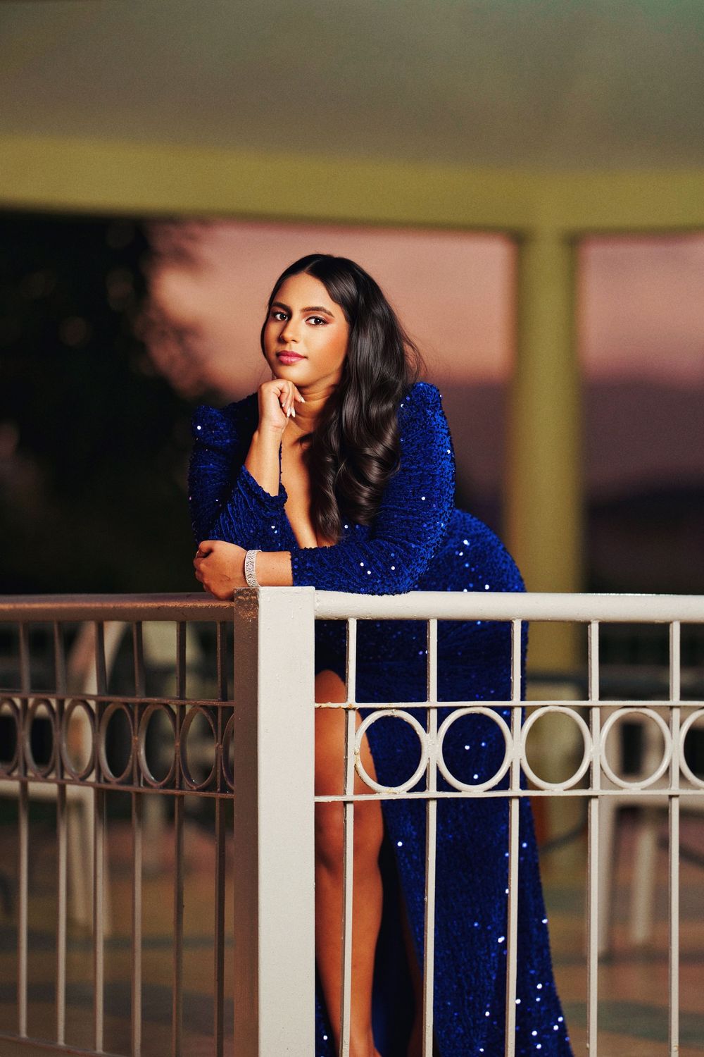 A person in a sparkling royal blue evening gown leans thoughtfully against a white railing at dusk.