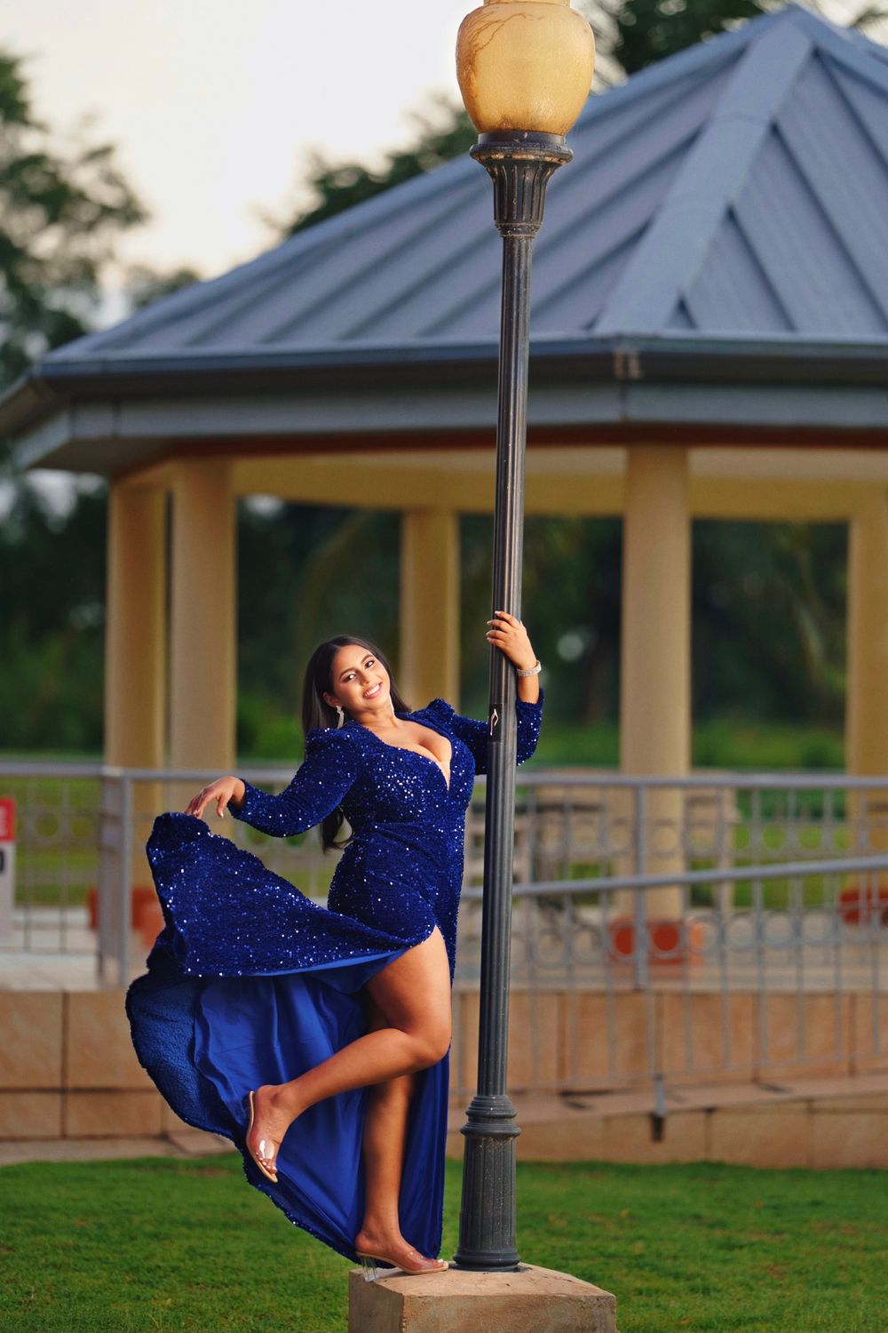 A person in a sparkling royal blue dress poses gracefully by a vintage lamp post near a gazebo at sunset.
