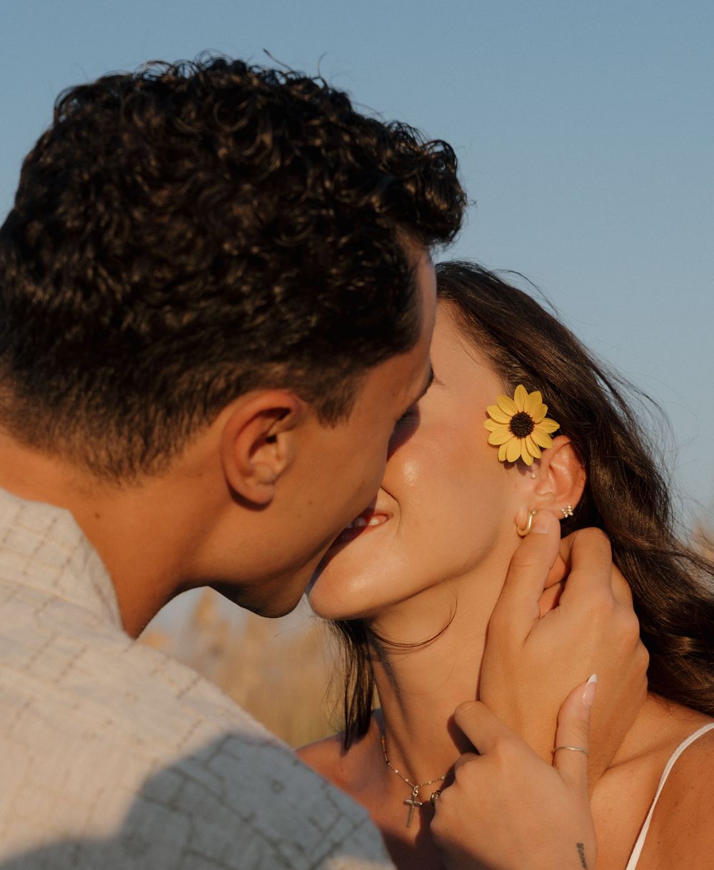 A romantic couple shares an intimate kiss at sunset with a sunflower in her hair.