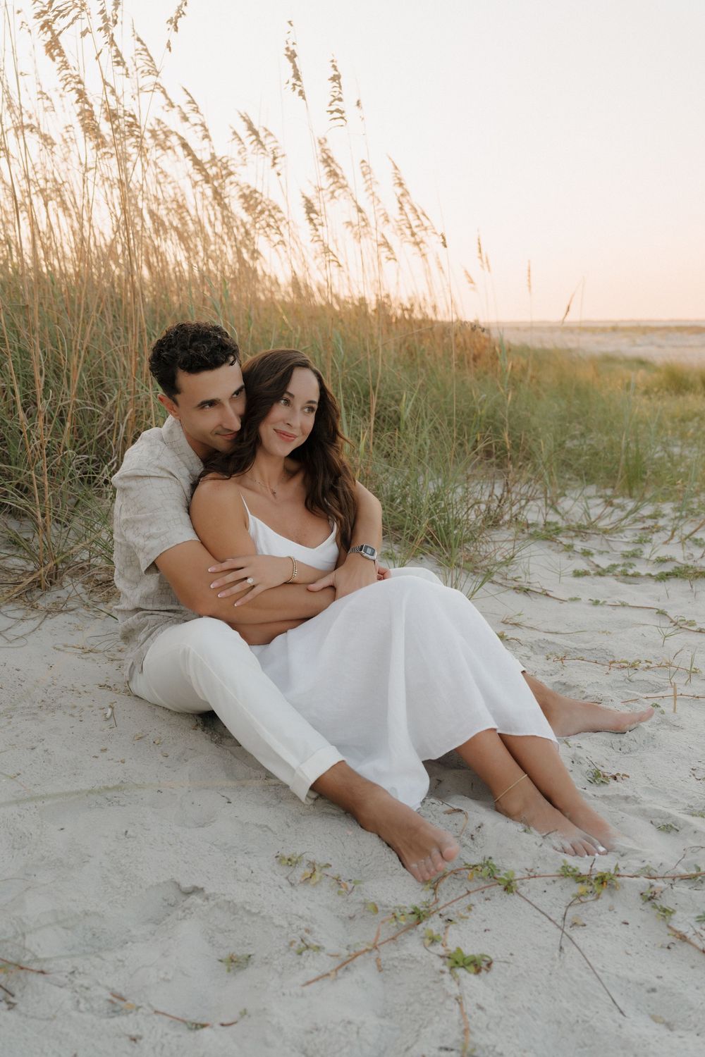 A romantic couple sits together on a sandy beach at sunset with tall grass swaying in the background.