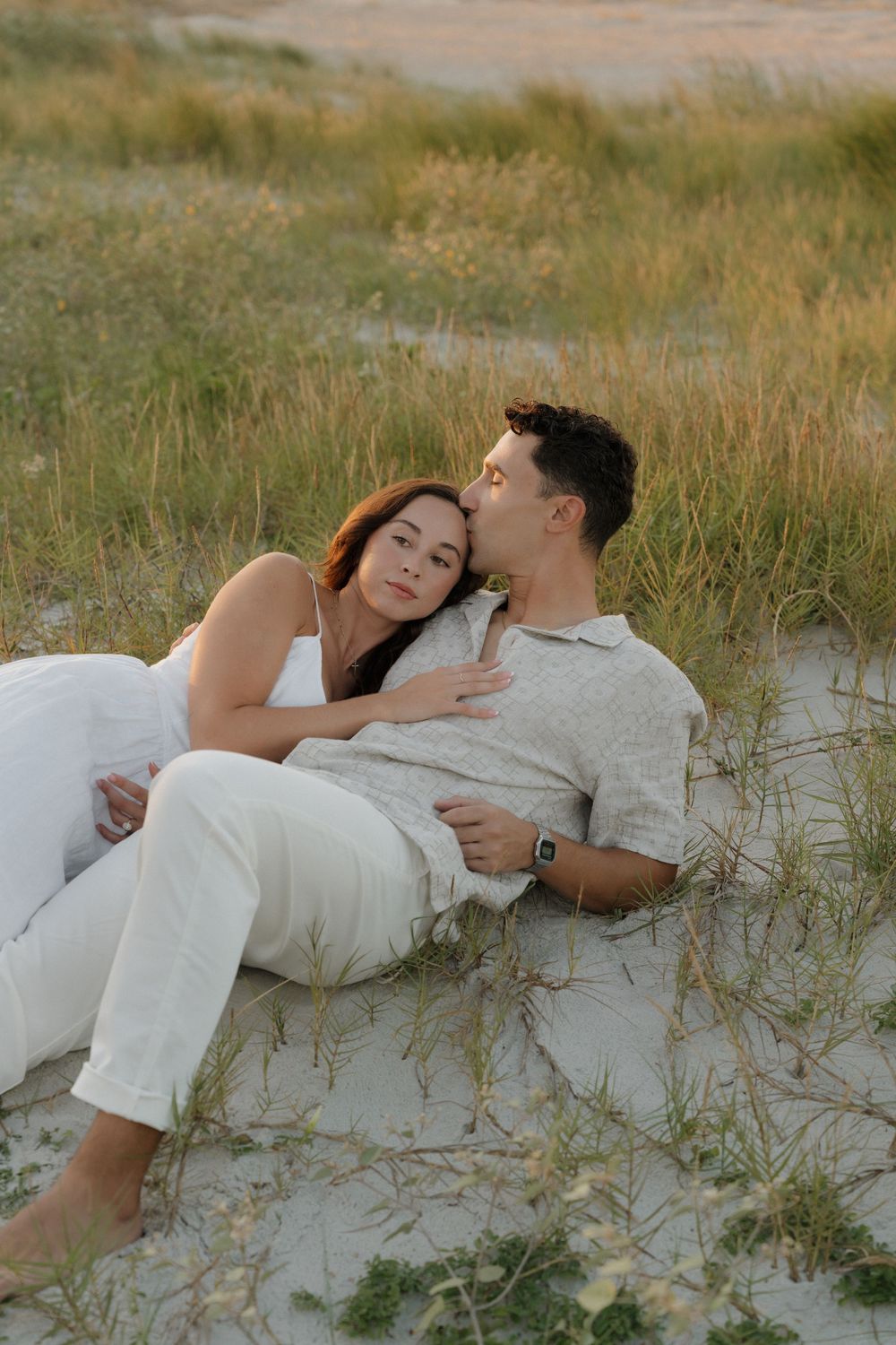A couple relaxes together on sandy dunes during a romantic beach photoshoot at sunset.