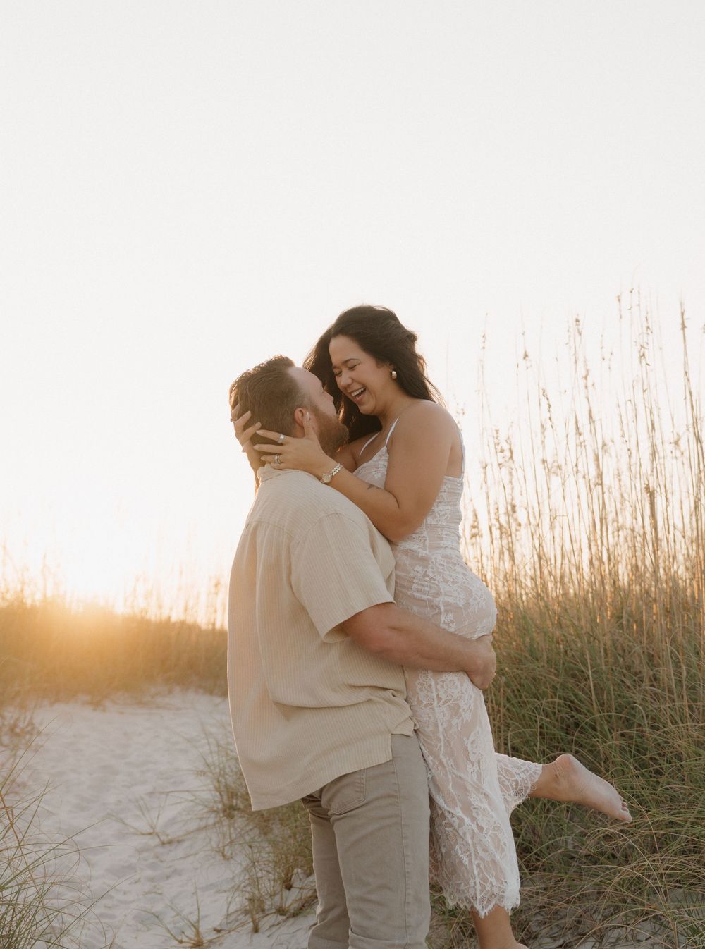 Couple embraces on sandy beach at sunset with tall grass swaying in the golden light.