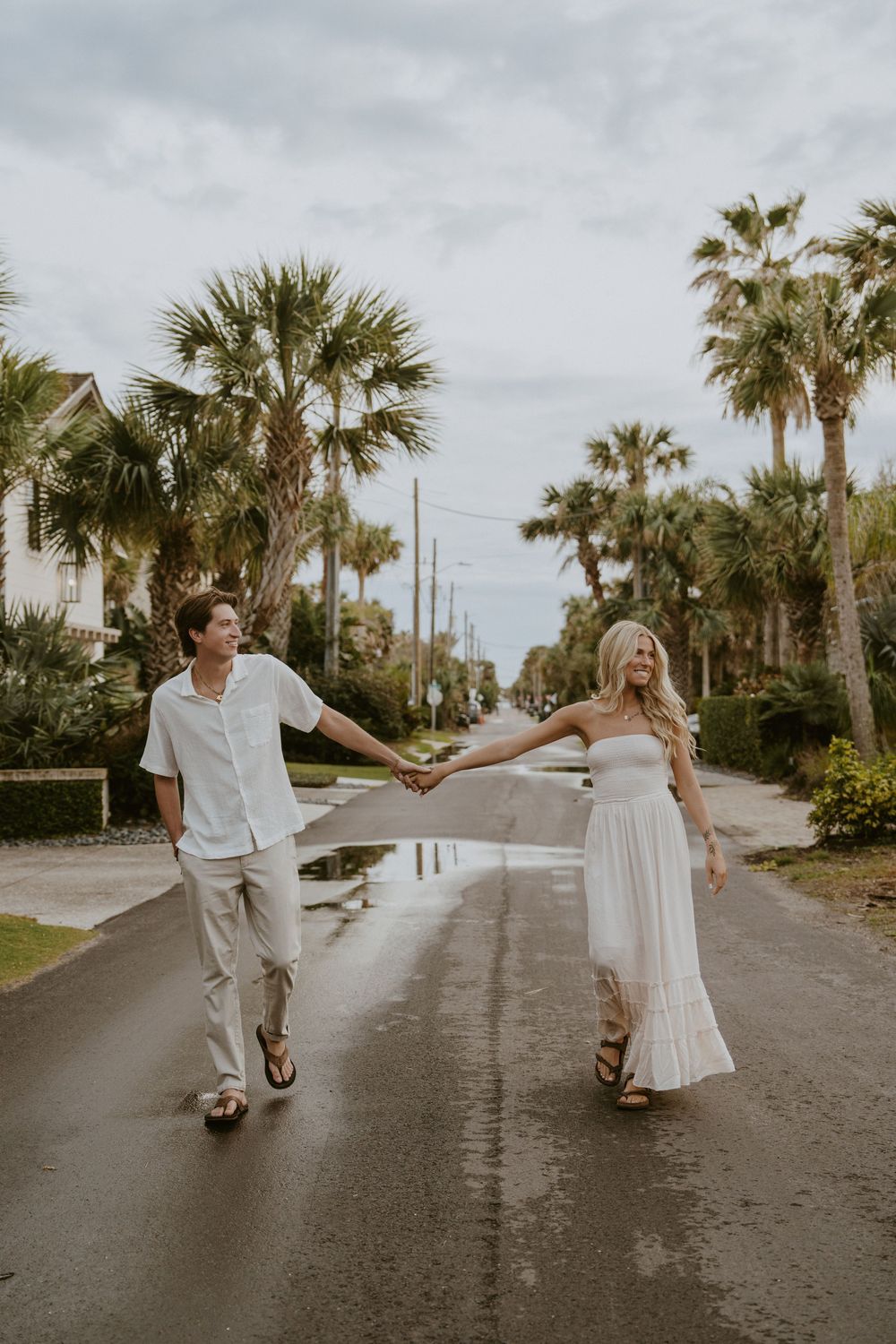 A couple in white outfits dances playfully on a palm tree-lined street near the beach.