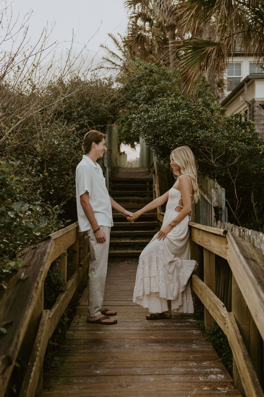 A couple holds hands on a wooden boardwalk surrounded by palm trees and coastal vegetation at sunset.