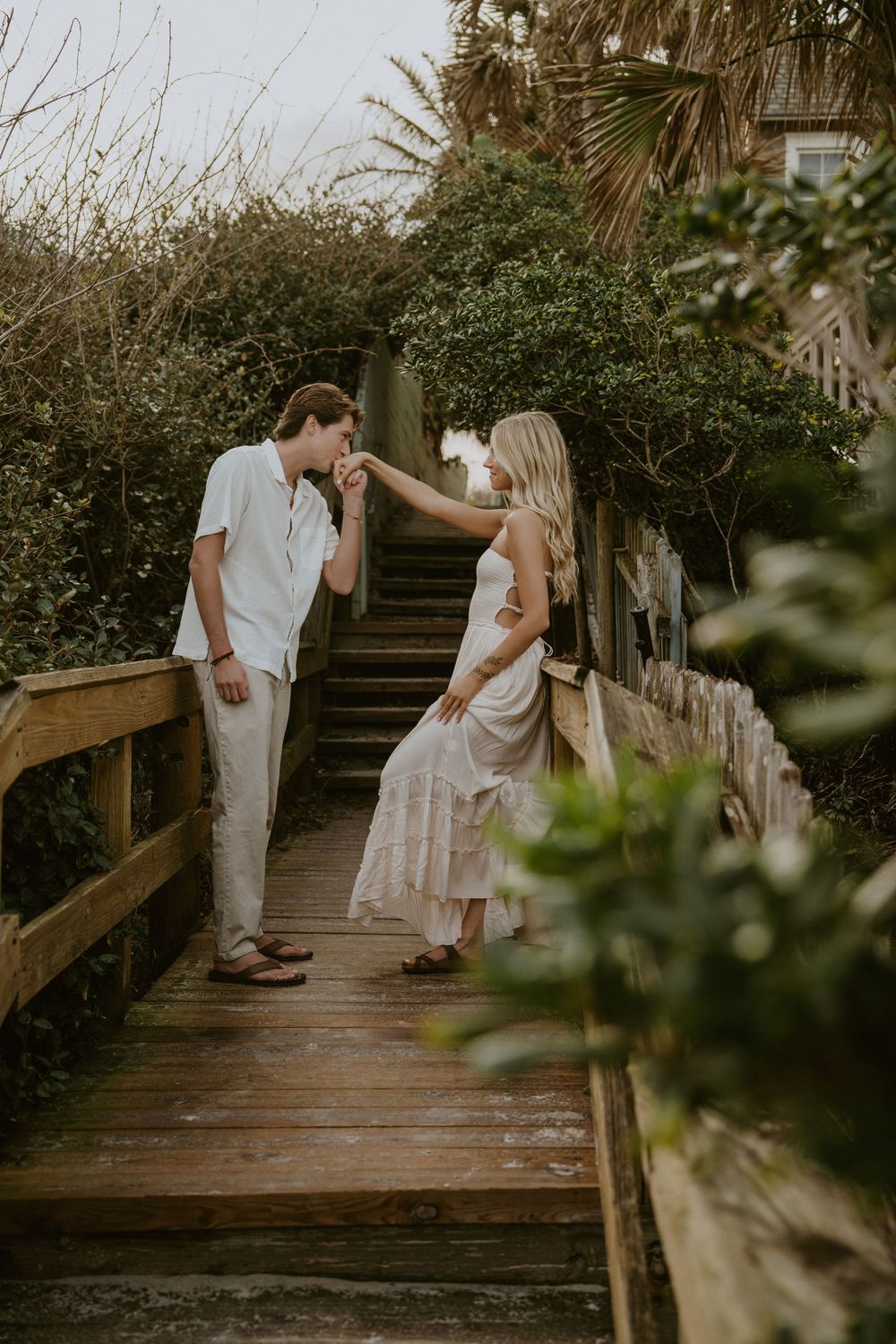 A couple shares sweet moments on wooden boardwalk steps surrounded by lush coastal vegetation.