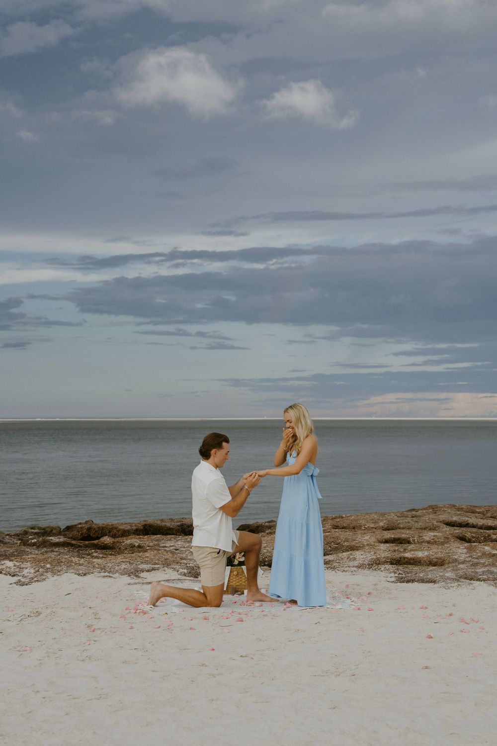 A romantic beach proposal scene at sunset with scattered rose petals on the sandy shore.
