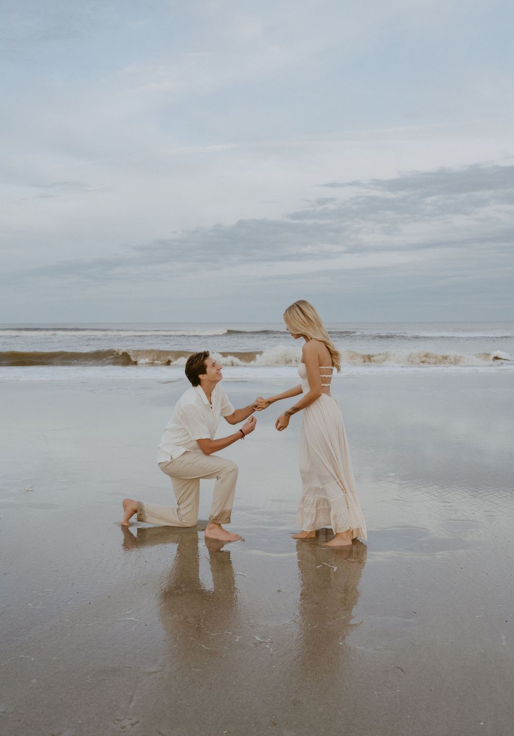 A romantic beach proposal scene at sunset with two people standing in shallow ocean water.