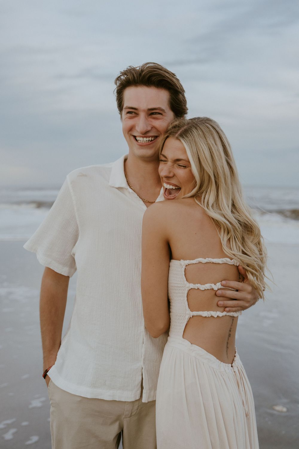 A romantic couple embraces on a cloudy beach, wearing white and beige clothing in a series of intimate poses.
