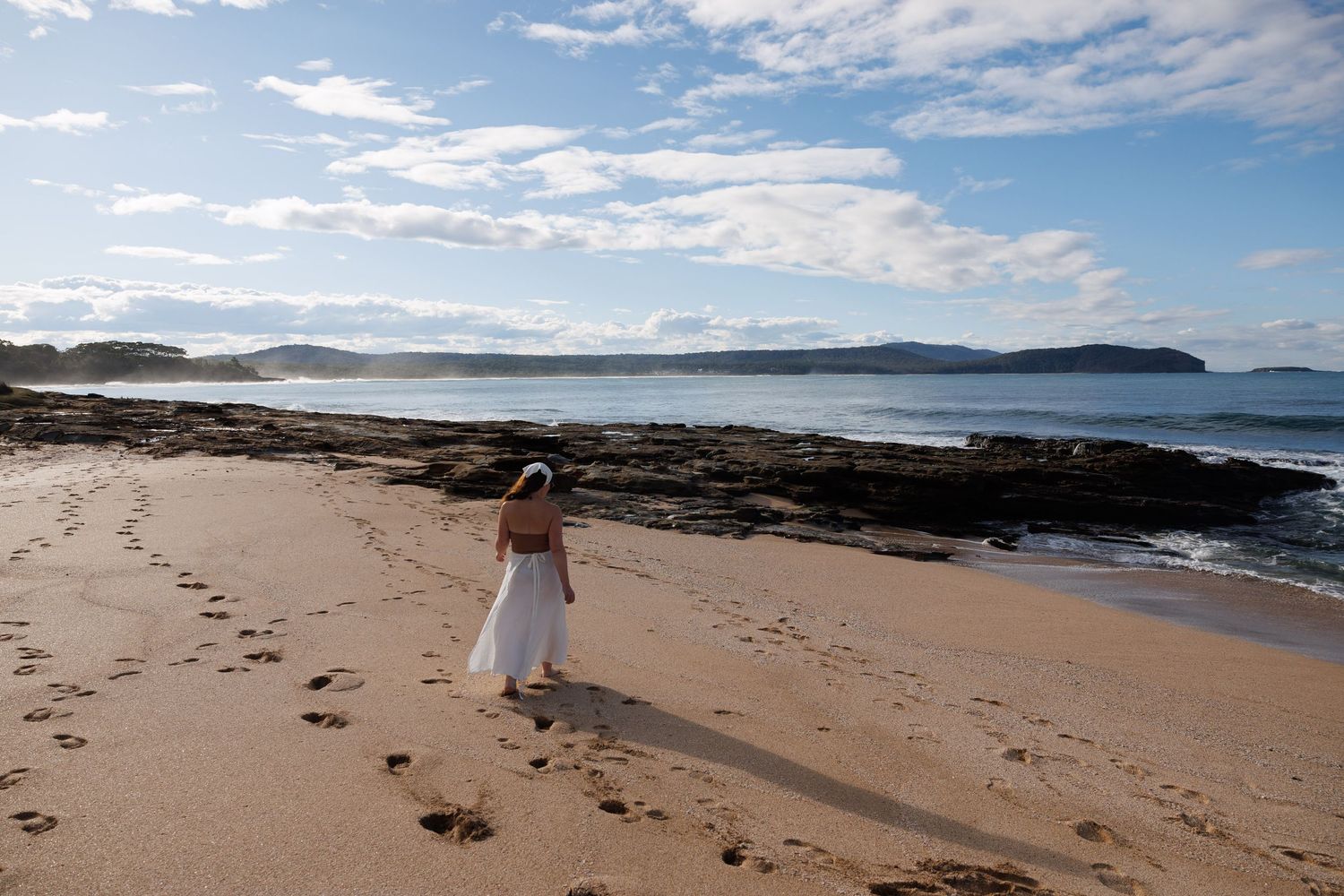 A beautiful coastal landscape with sandy beach, rocky outcrops, and blue ocean waves under a cloudy sky.
