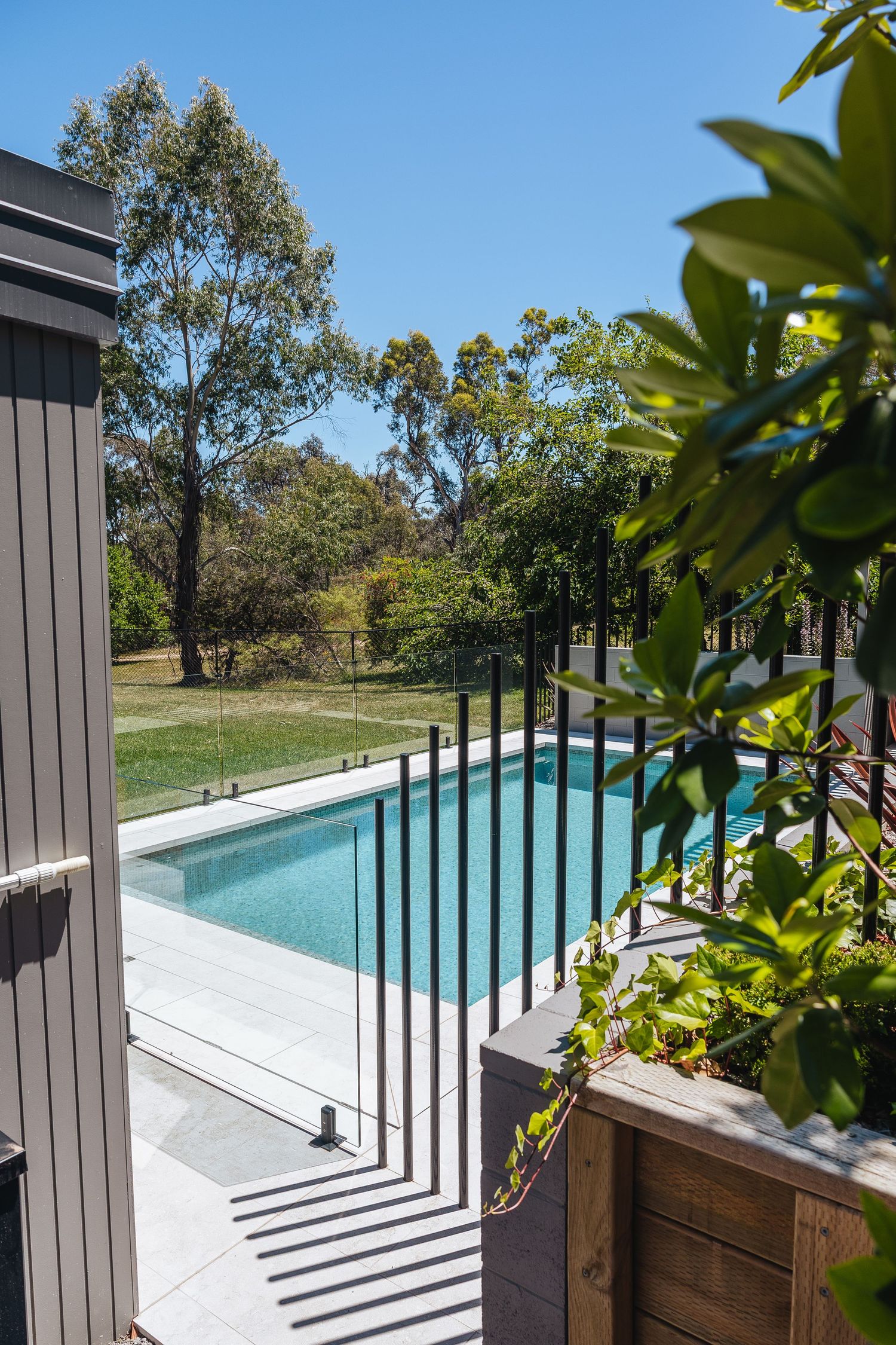 Modern outdoor swimming pool with glass fence panels and wooden decking surrounded by lush green landscaping.