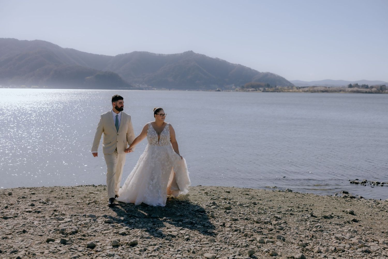A couple in wedding attire walks hand in hand along a scenic beach with mountains and sparkling water in the background.