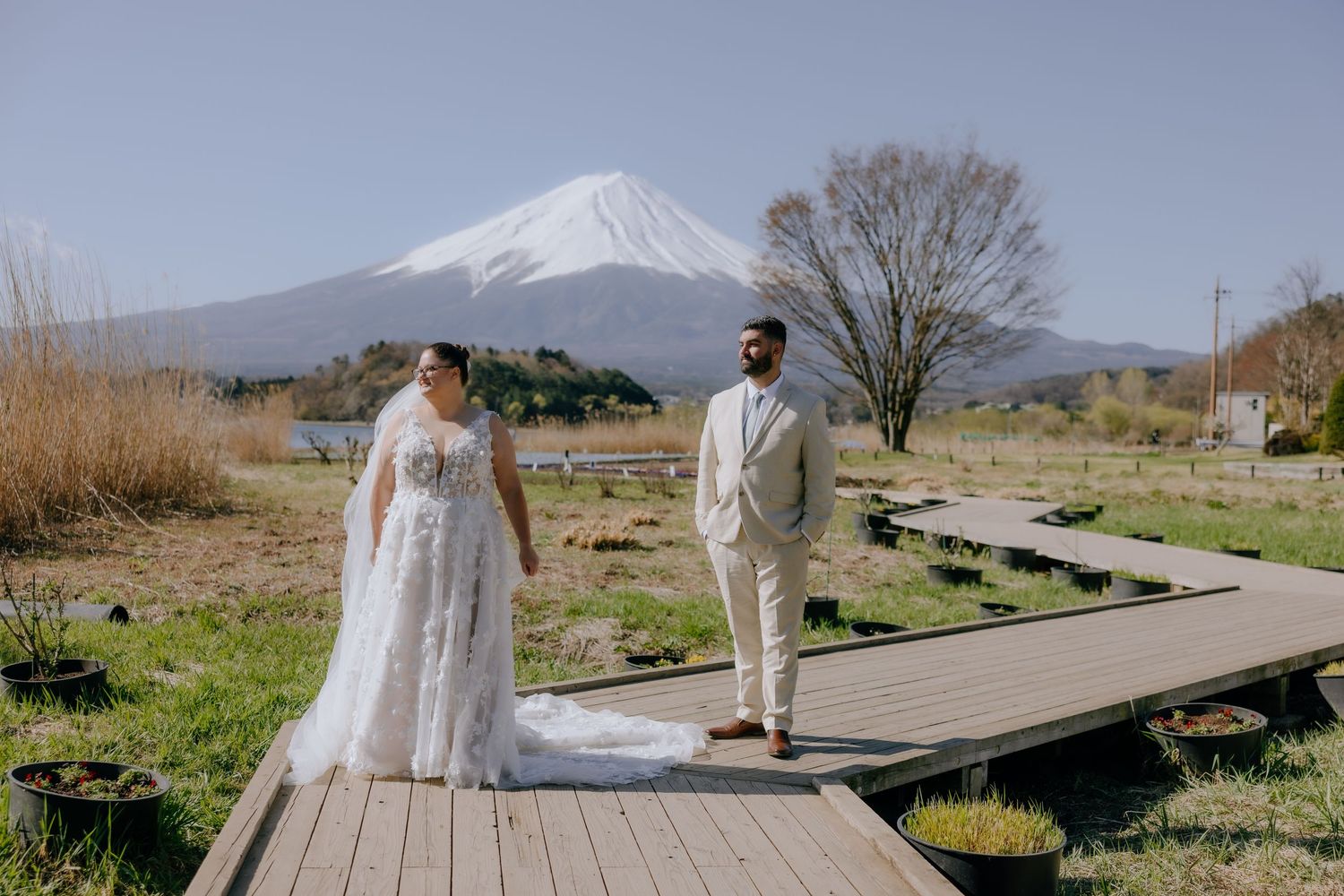 Newlyweds walk along a scenic lakeside path with snow-capped Mount Fuji in the background.