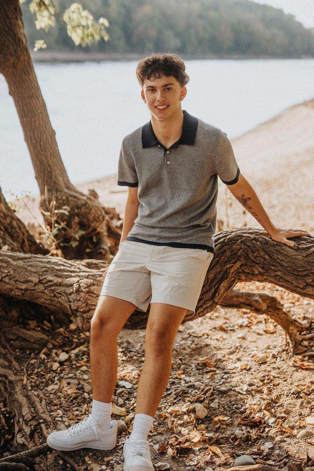 Person wearing grey polo shirt and white shorts sits on fallen tree trunk by lakeside beach.