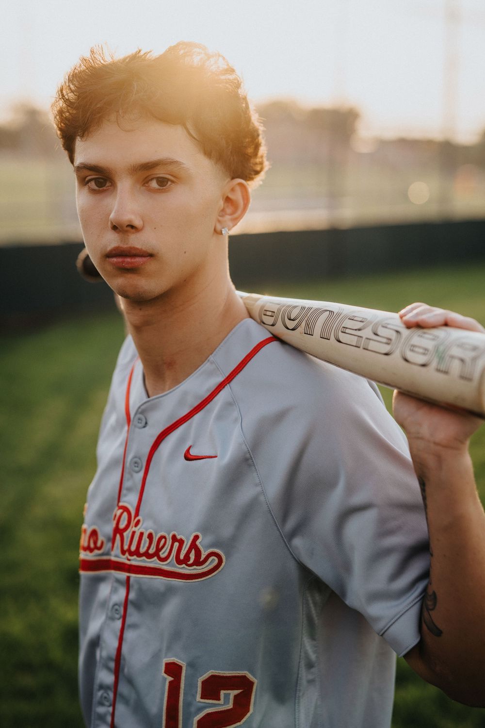 Baseball player in grey Two Rivers jersey holding bat against sunset background.