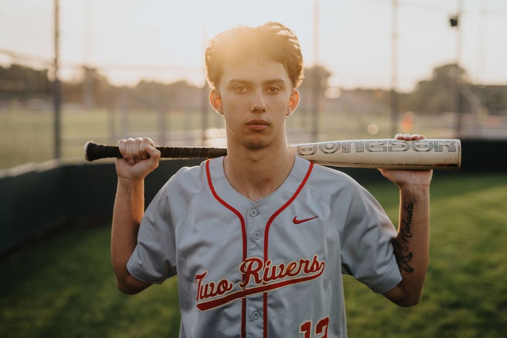 Baseball player in gray jersey with red lettering holds bat across shoulders at sunset on baseball field.