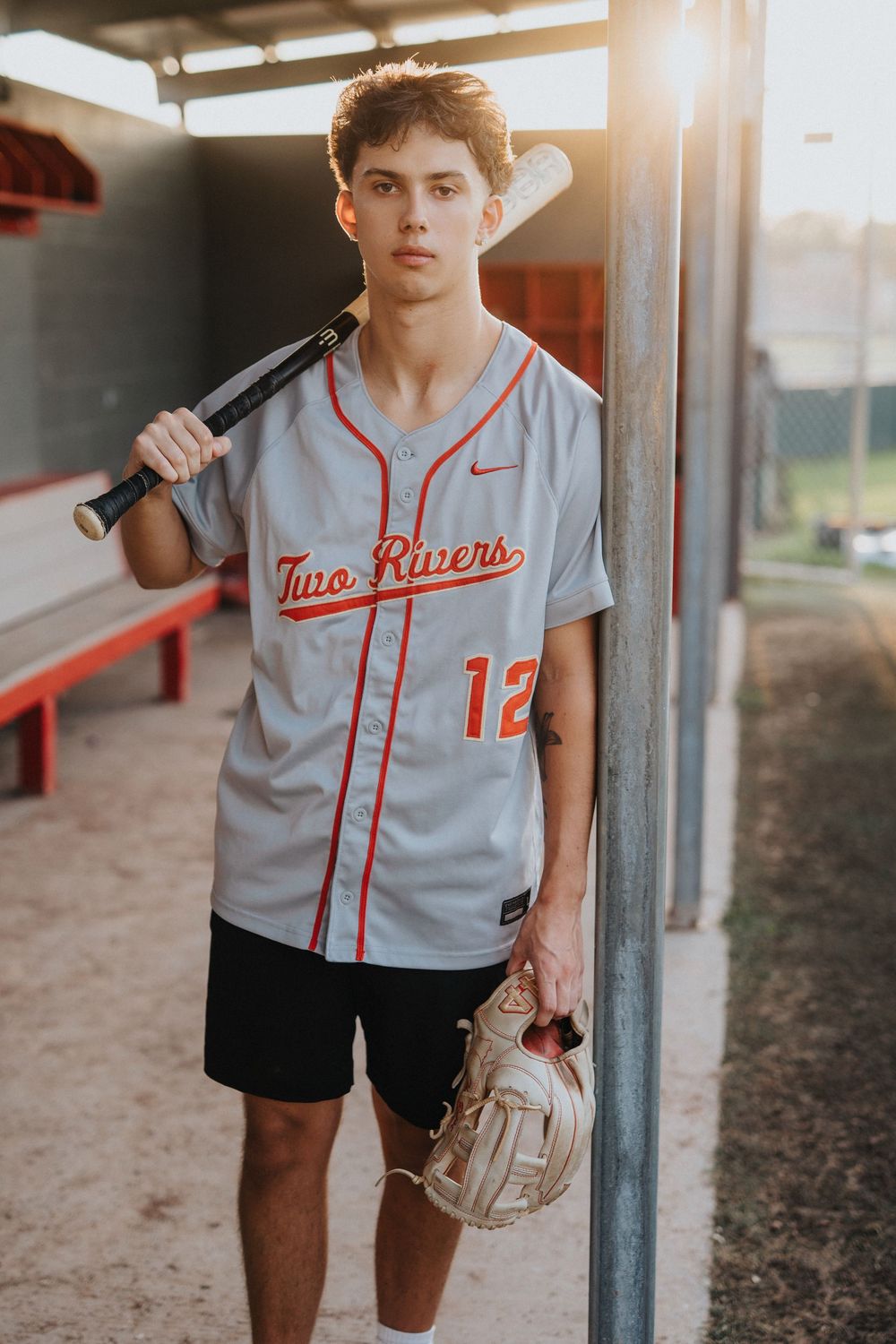 Youth baseball player in grey uniform with number 12 standing near dugout.