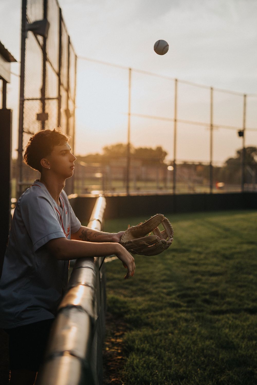 Softball player leaning on dugout fence at sunset while practicing catching drills in golden light.