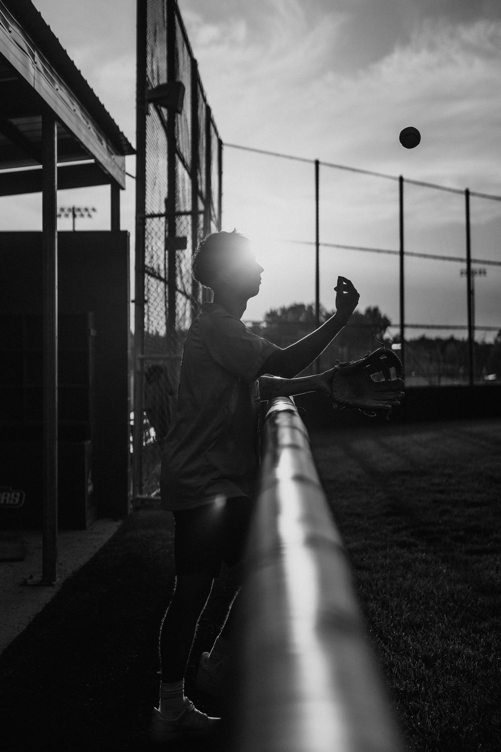 Silhouetted baseball player practices against chain link fence at dusk in black and white.