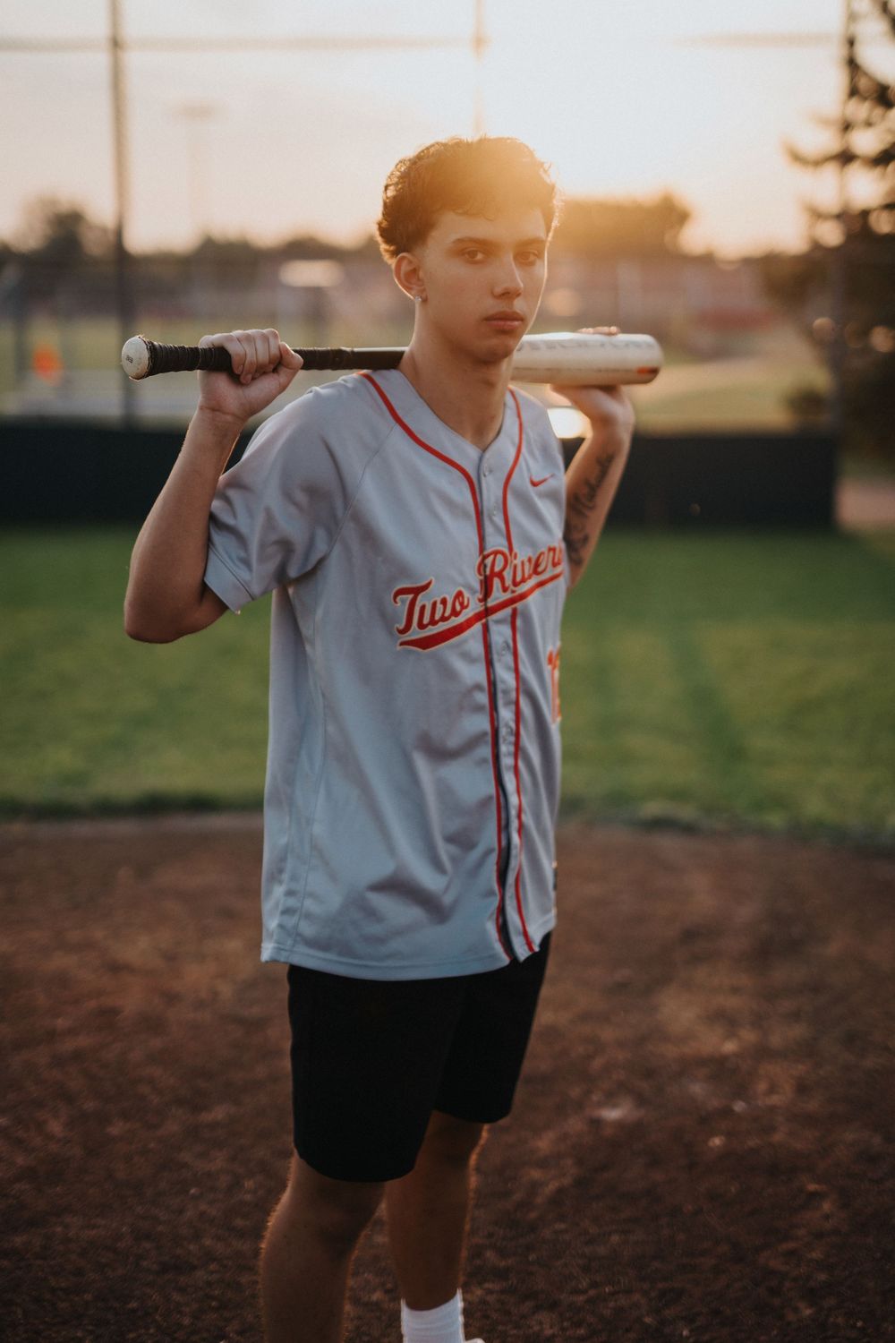 A baseball player in a white jersey with 'Two' written on it holds a bat at sunset on a dirt field.