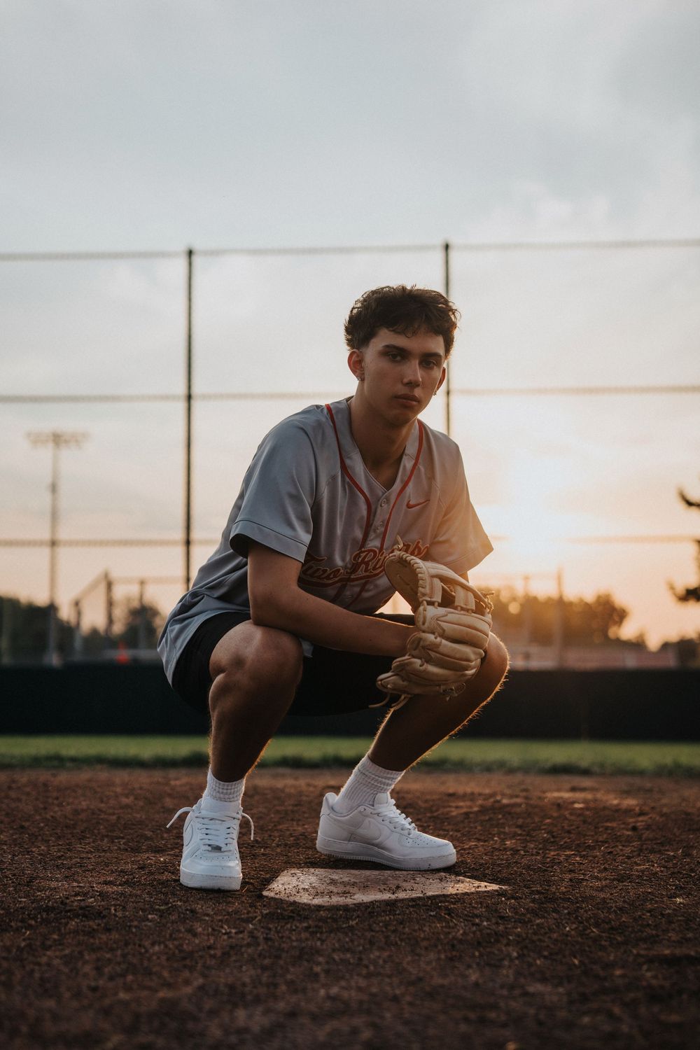 Baseball player crouching at base during golden sunset on dirt field wearing white sneakers and denim.