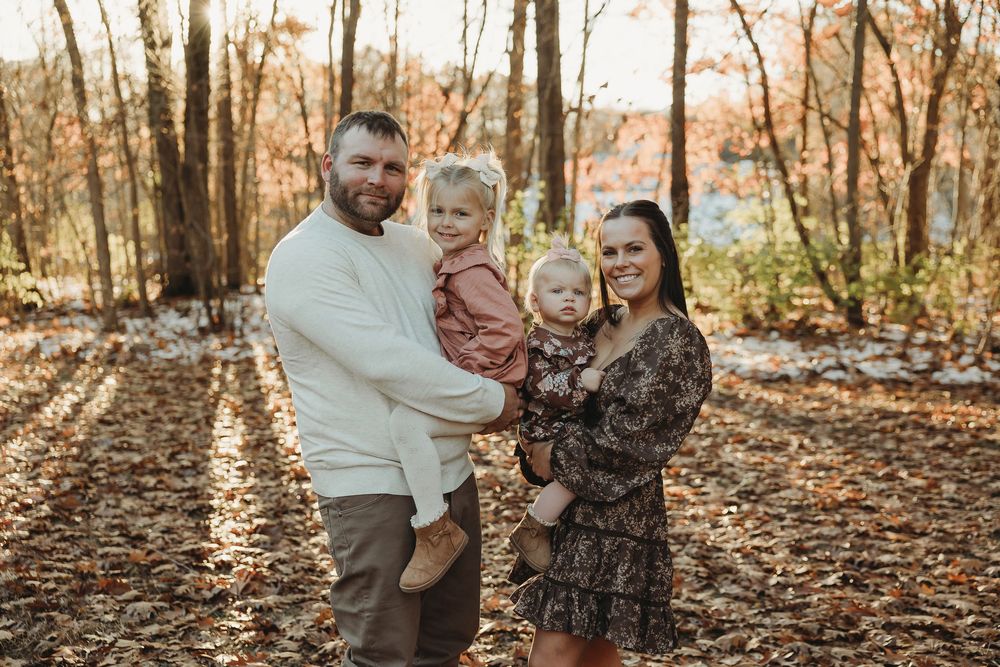 A family enjoys a tender moment together while walking through a forest covered in fallen autumn leaves.