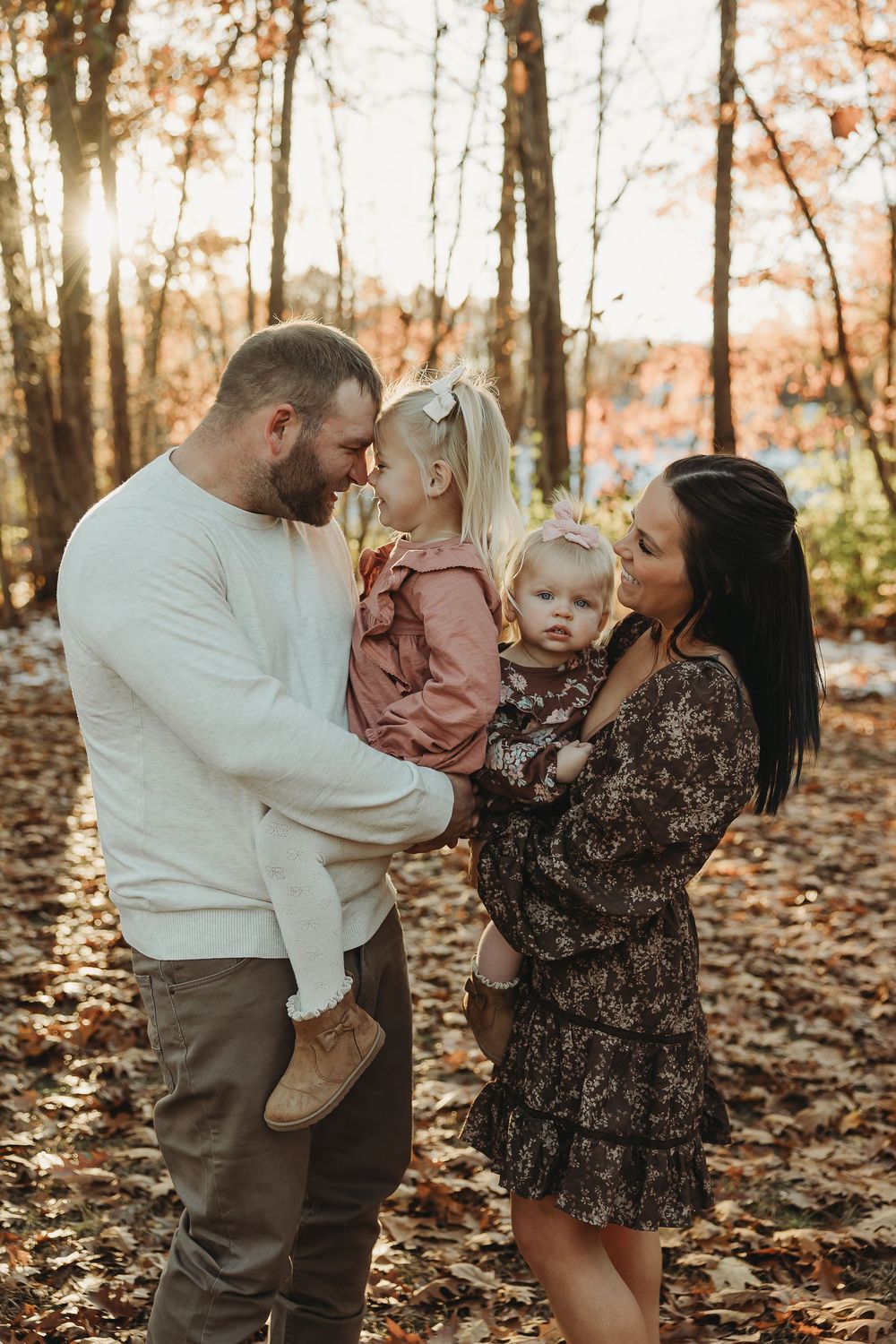 A couple and their toddler share tender moments in a fall forest setting with golden leaves scattered on the ground.