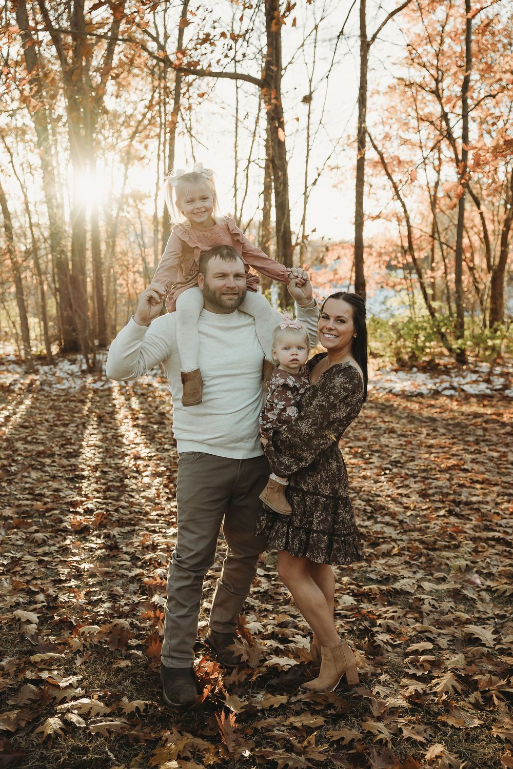 A couple and child play together in a sunlit autumn forest with fallen leaves covering the ground.