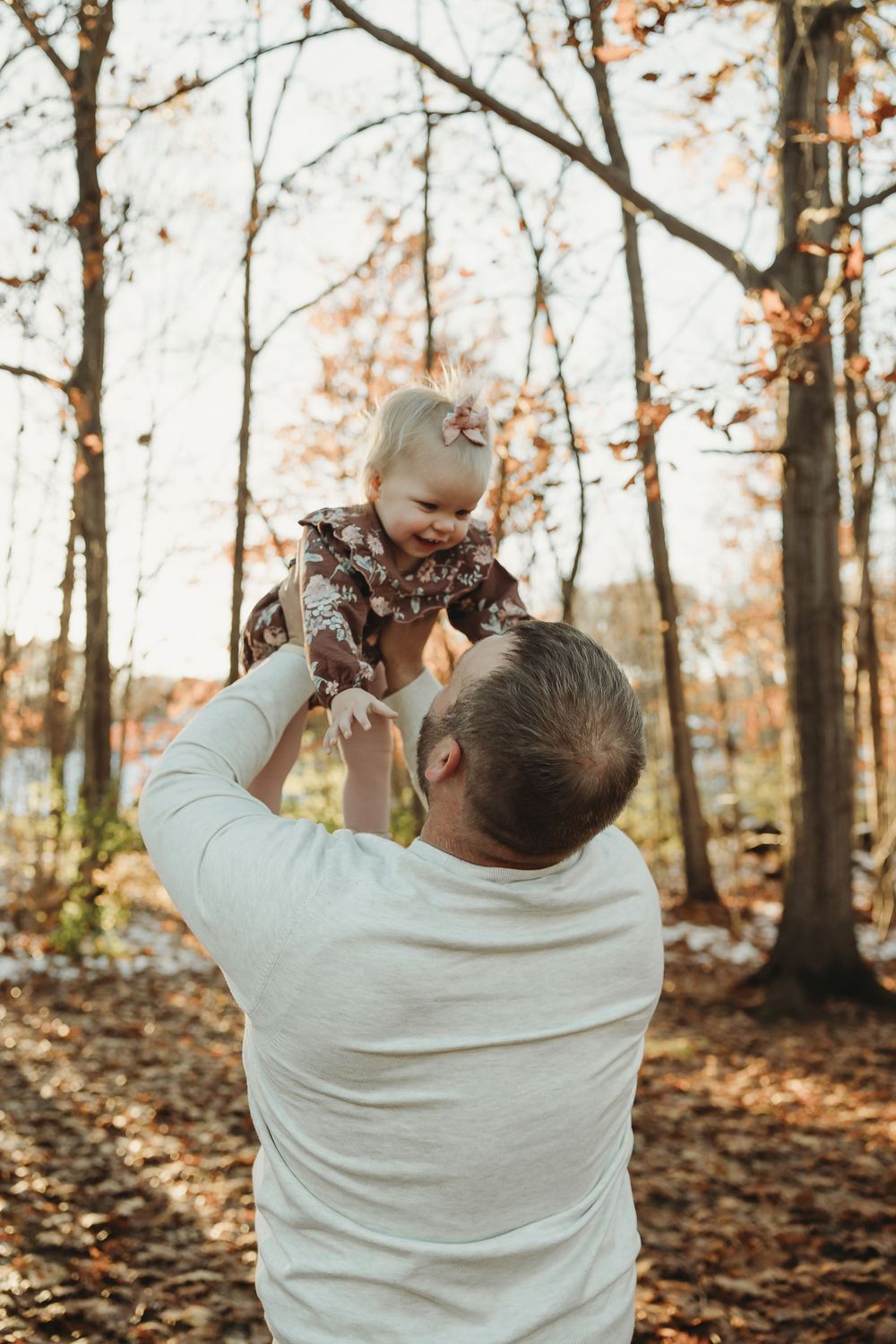 A tender moment captured in an autumn forest as a person in a white sweater lifts a small child wearing a floral outfit.