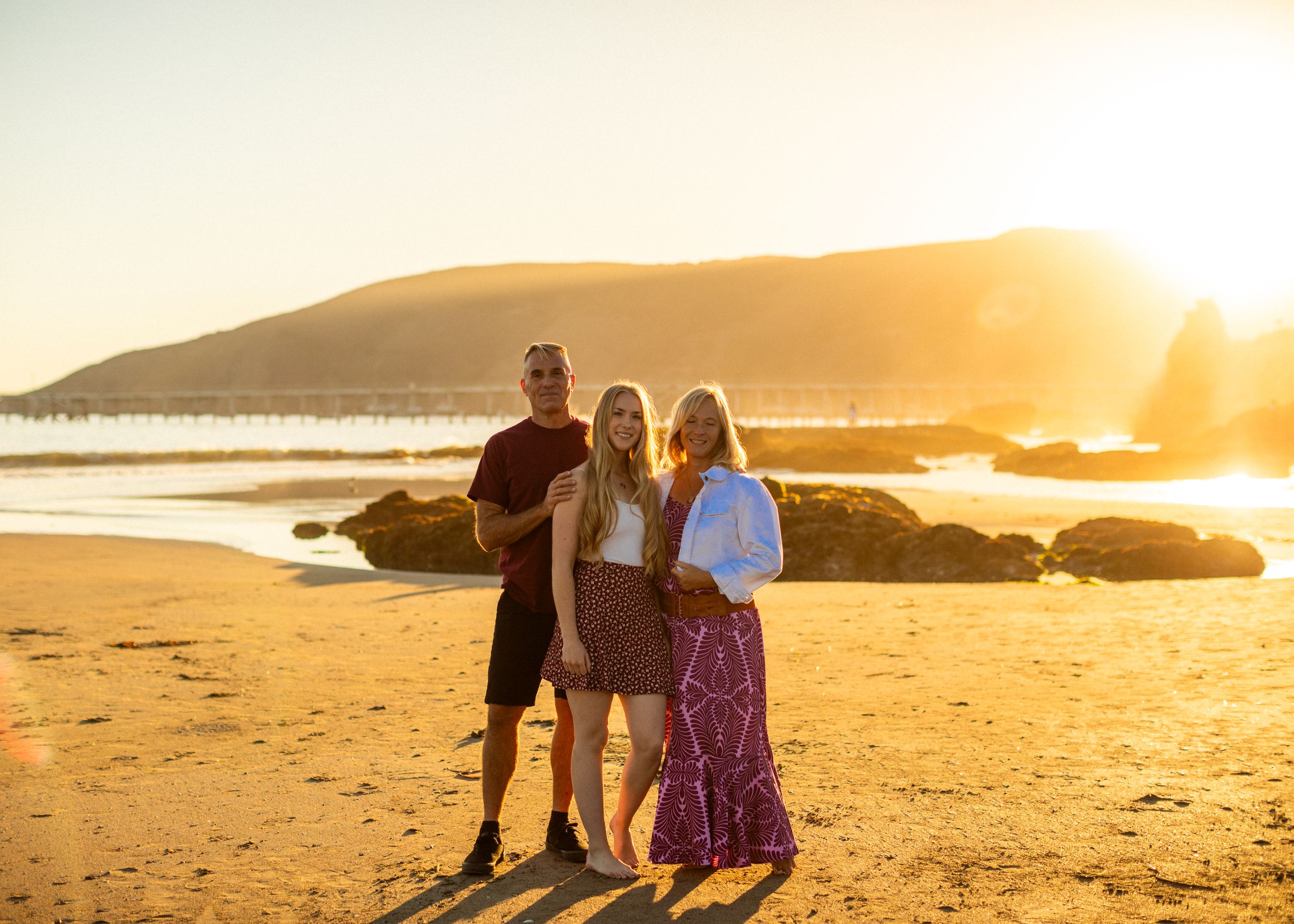 A small group poses on a beach at sunset with rocks and mountains in the background.