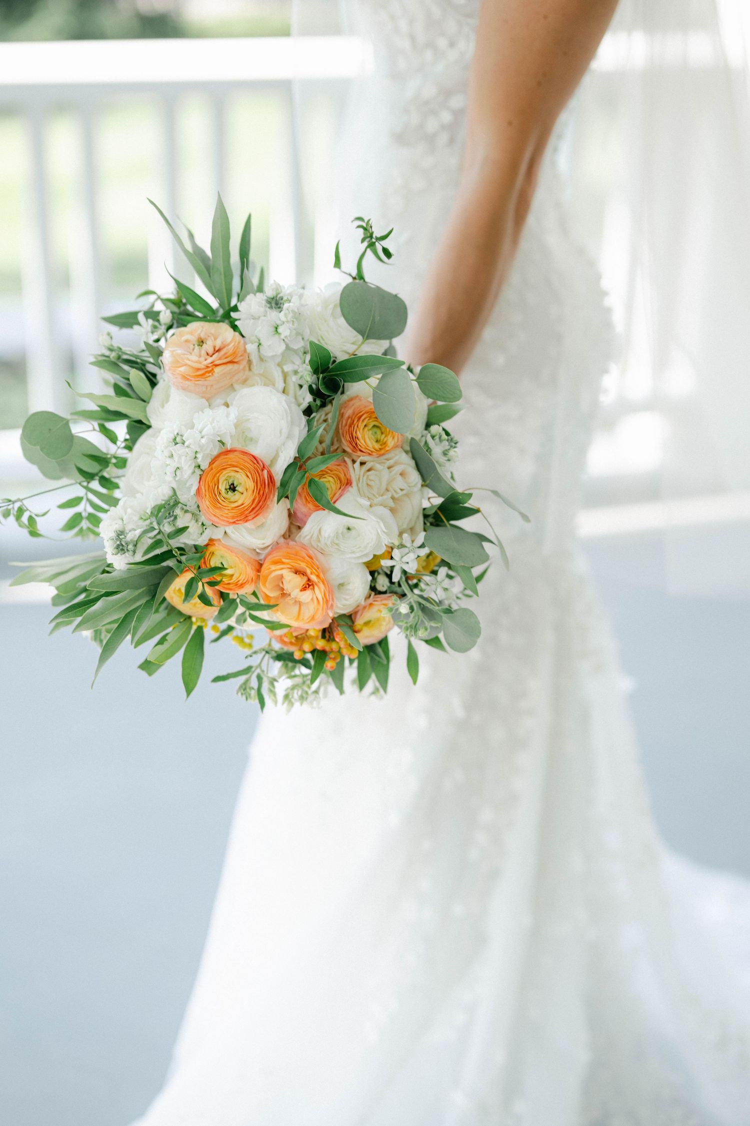 A bride in a flowing lace wedding gown with long veil holds a colorful bouquet while standing on a white porch. - Lake Forest