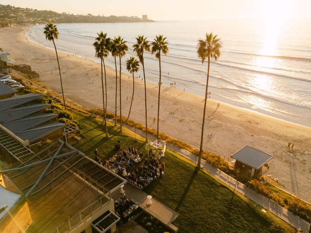 Aerial panoramic view of a beach with palm trees and golden sunlight reflecting off the ocean at sunset.