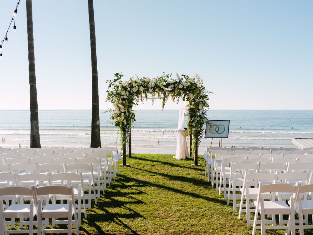 White wedding chairs arranged on a grassy oceanfront lawn with palm trees and a floral arch for a beach ceremony.