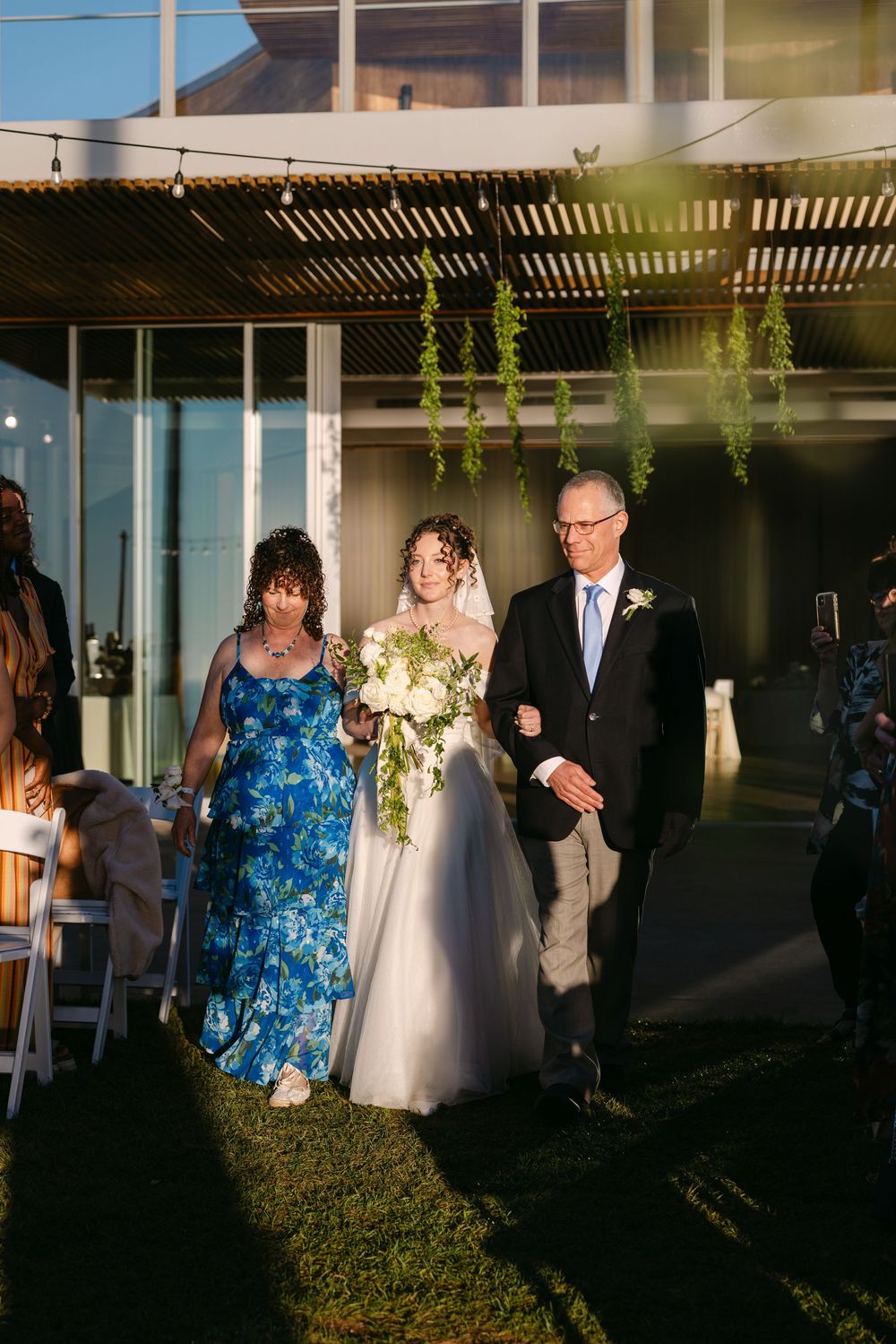 Wedding processional walking down an outdoor aisle under a pergola at sunset with guests watching.