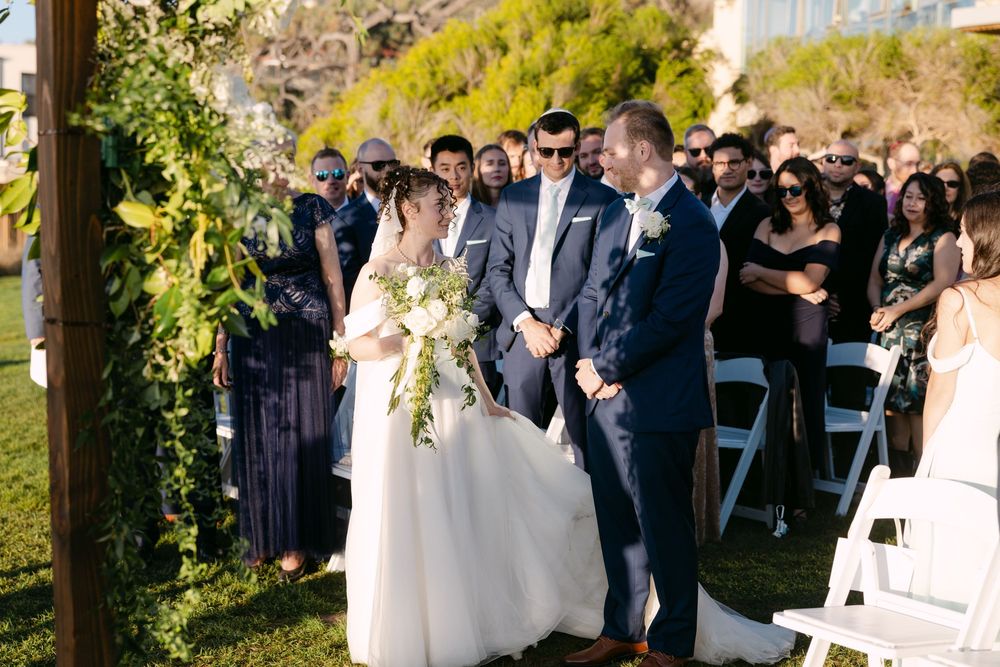 Outdoor wedding ceremony takes place under a wooden arch decorated with greenery and white flowers.