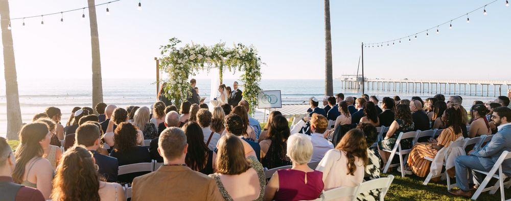 Wedding guests seated outdoors at a beachfront ceremony with string lights overhead and an ocean pier in the background.