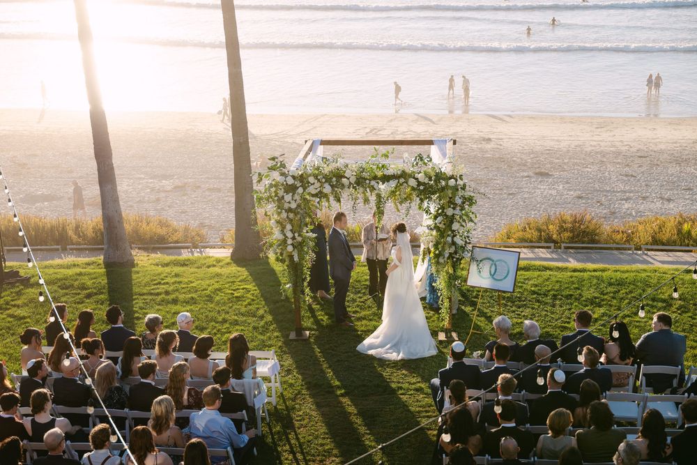 A scenic outdoor wedding ceremony takes place on a grassy lawn overlooking the ocean at sunset.