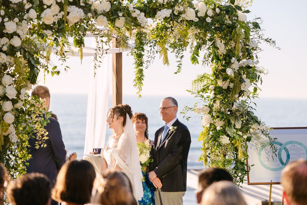 Outdoor wedding ceremony setup with white floral arch and chairs overlooking ocean at sunset.